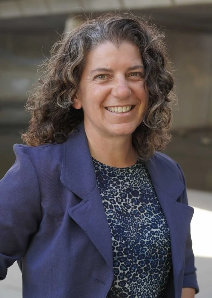 A woman with curly brown hair wearing a blue blazer and a leopard print top, smiling outdoors.