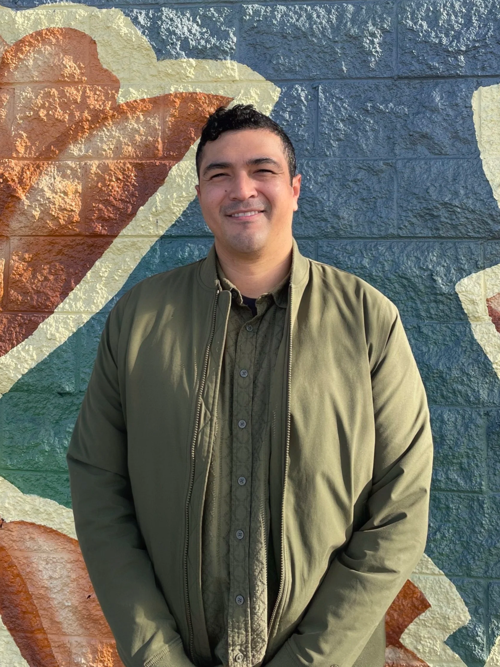 A man standing in front of a colorful mural painted on a brick wall, smiling at the camera, wearing an olive green jacket and shirt.