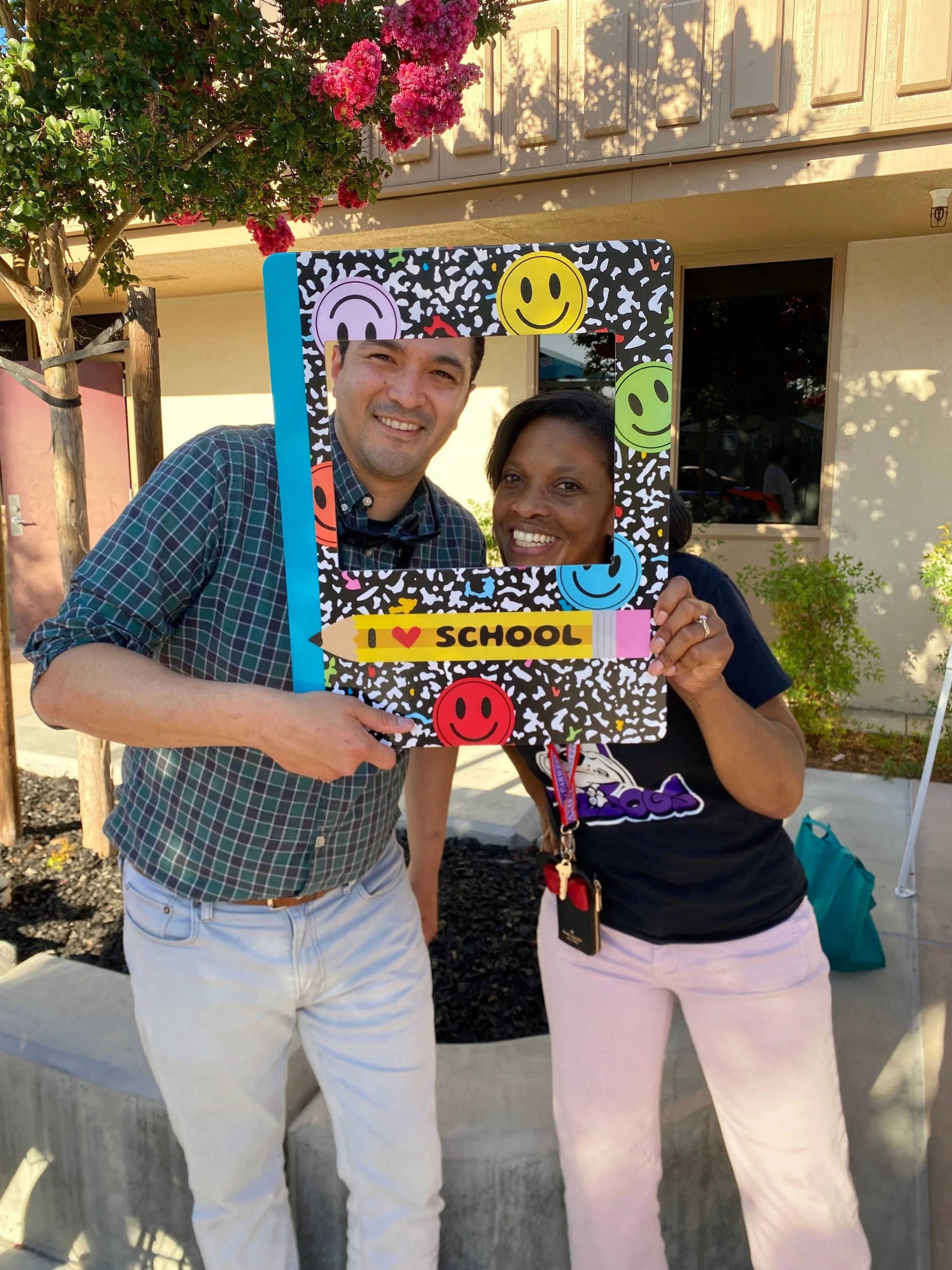 Two smiling adults standing outdoors at a school event, holding a colorful photo frame with smiley face decorations that says "I ❤️ SCHOOL".