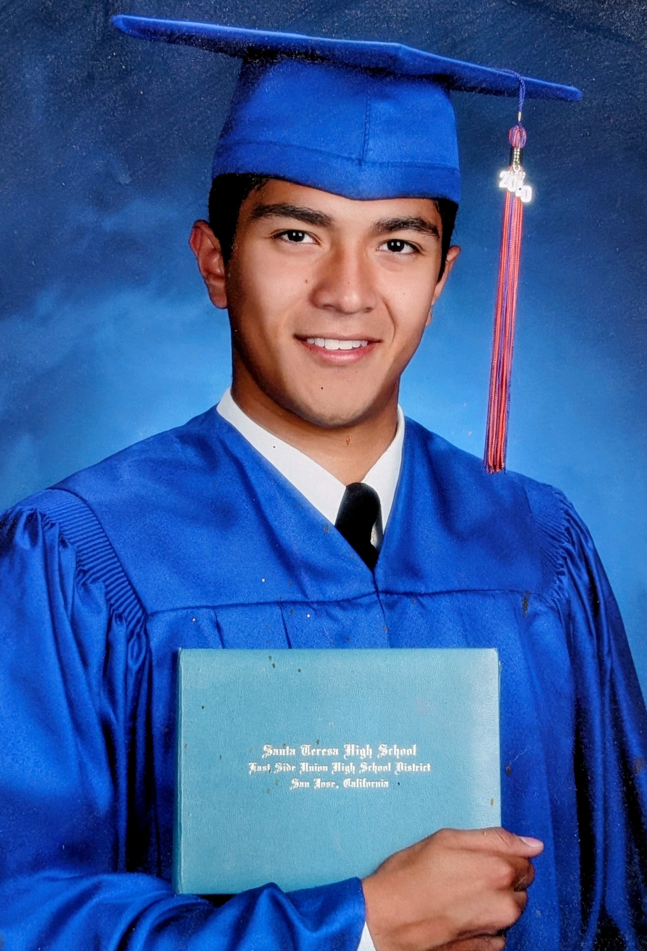 A young man in a blue graduation gown and cap, holding a diploma from Santa Teresa High School in San Jose, California.