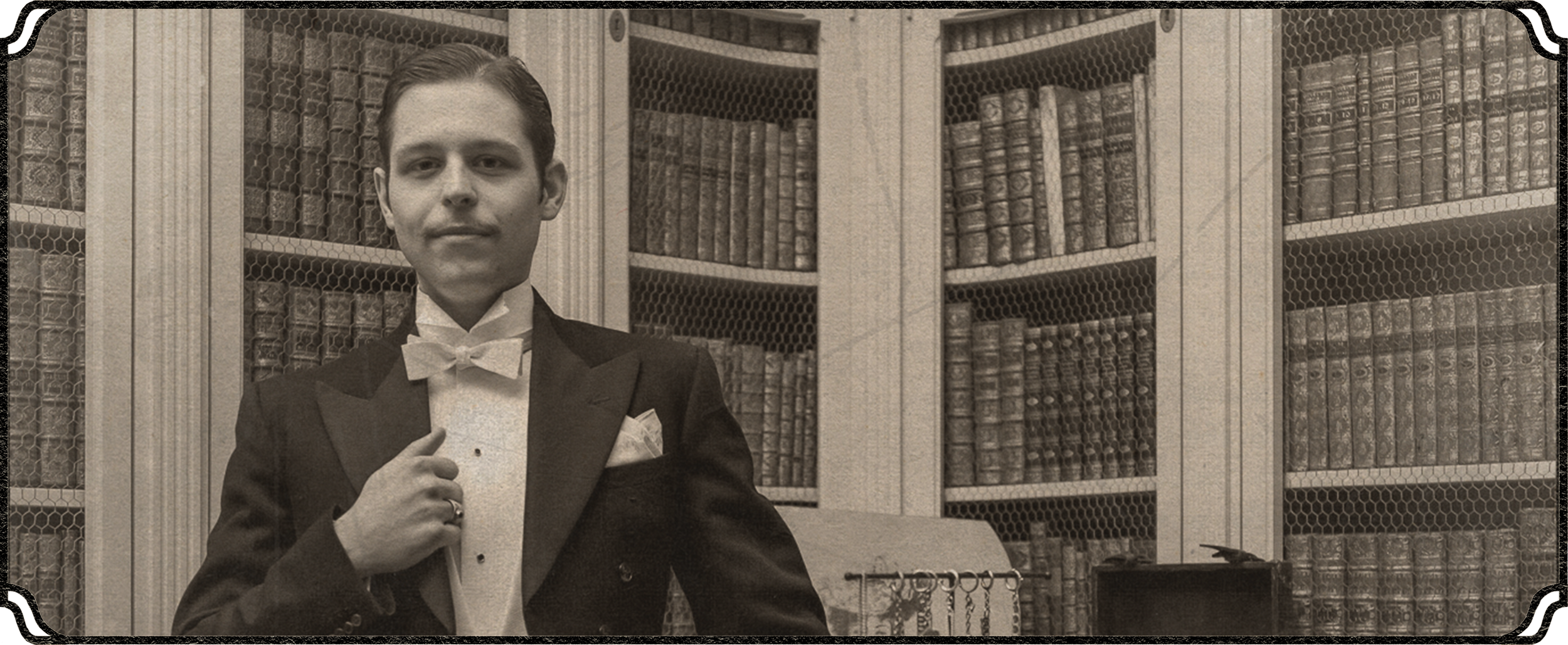 A young man dressed in a tuxedo and bow tie, standing next to a bookshelf filled with leather-bound books, in black and white.