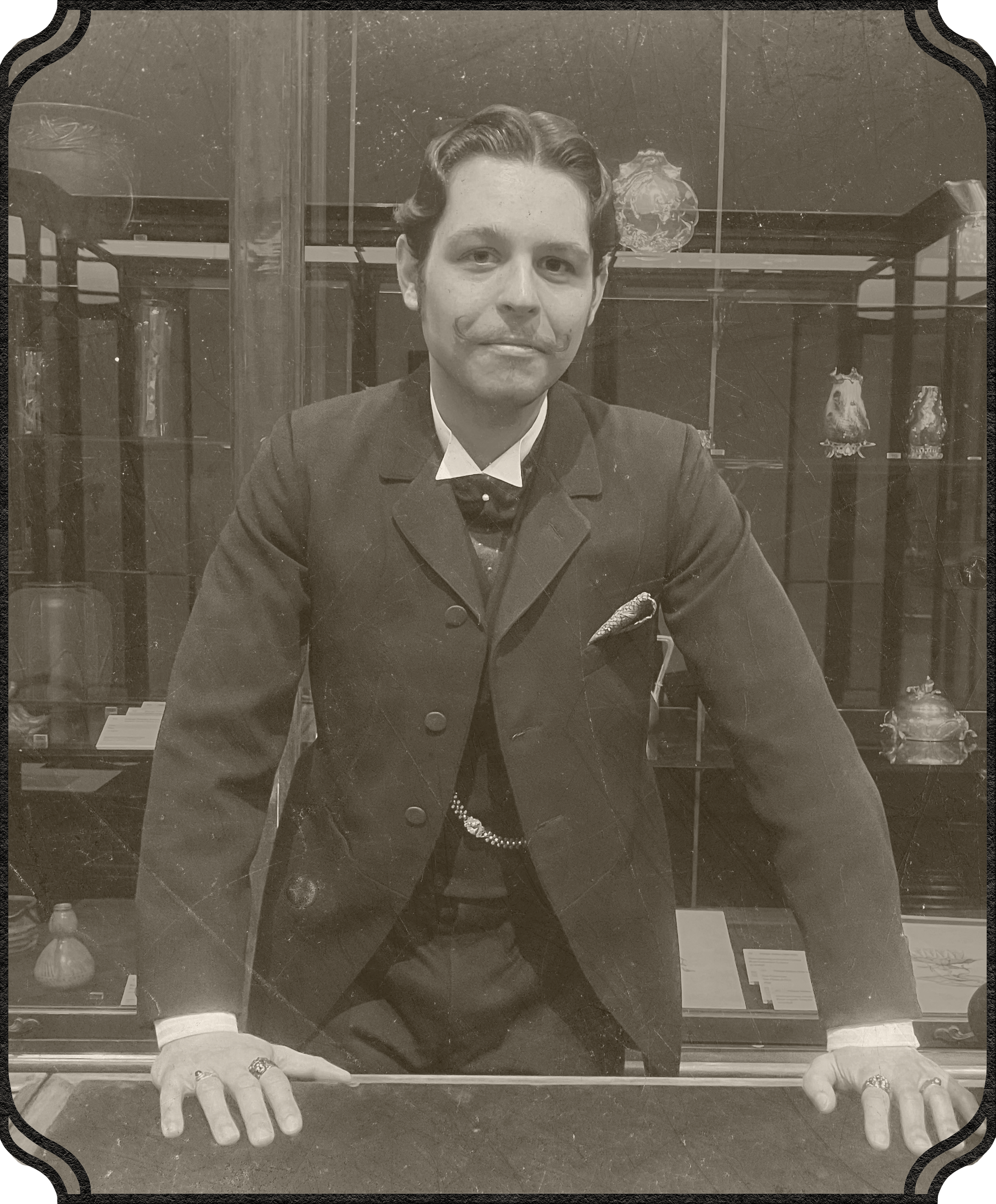 A man in a dark coat with a white collar and bow tie is looking through a glass display case at various items inside, including jewelry and decorative objects.