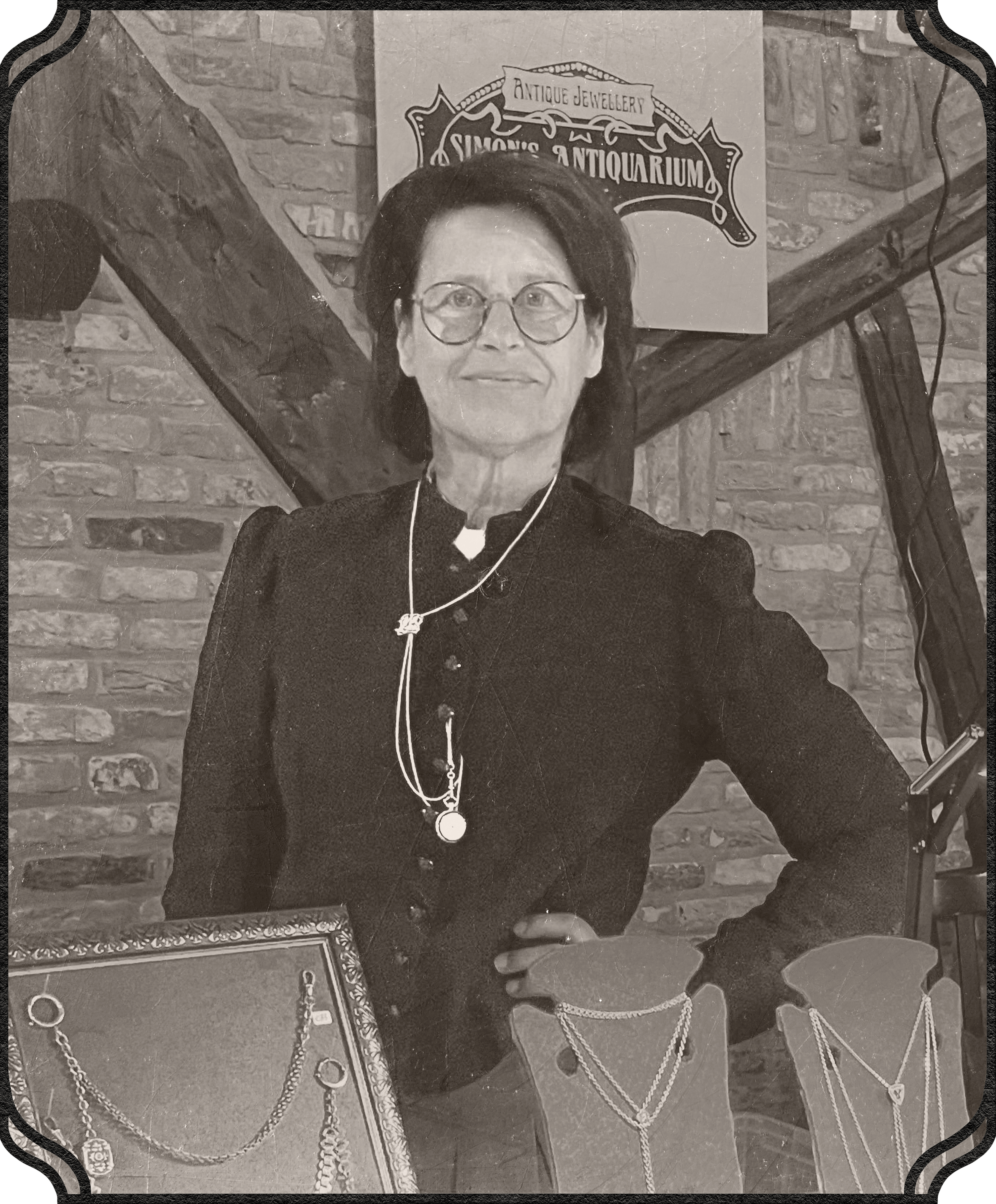 A woman wearing glasses, jewelry, and a dark dress stands indoors in front of a brick wall with a sign that reads 'Antique Jewelry Simon's Antiquarium.' She is looking at the camera and has a pleasant expression.
