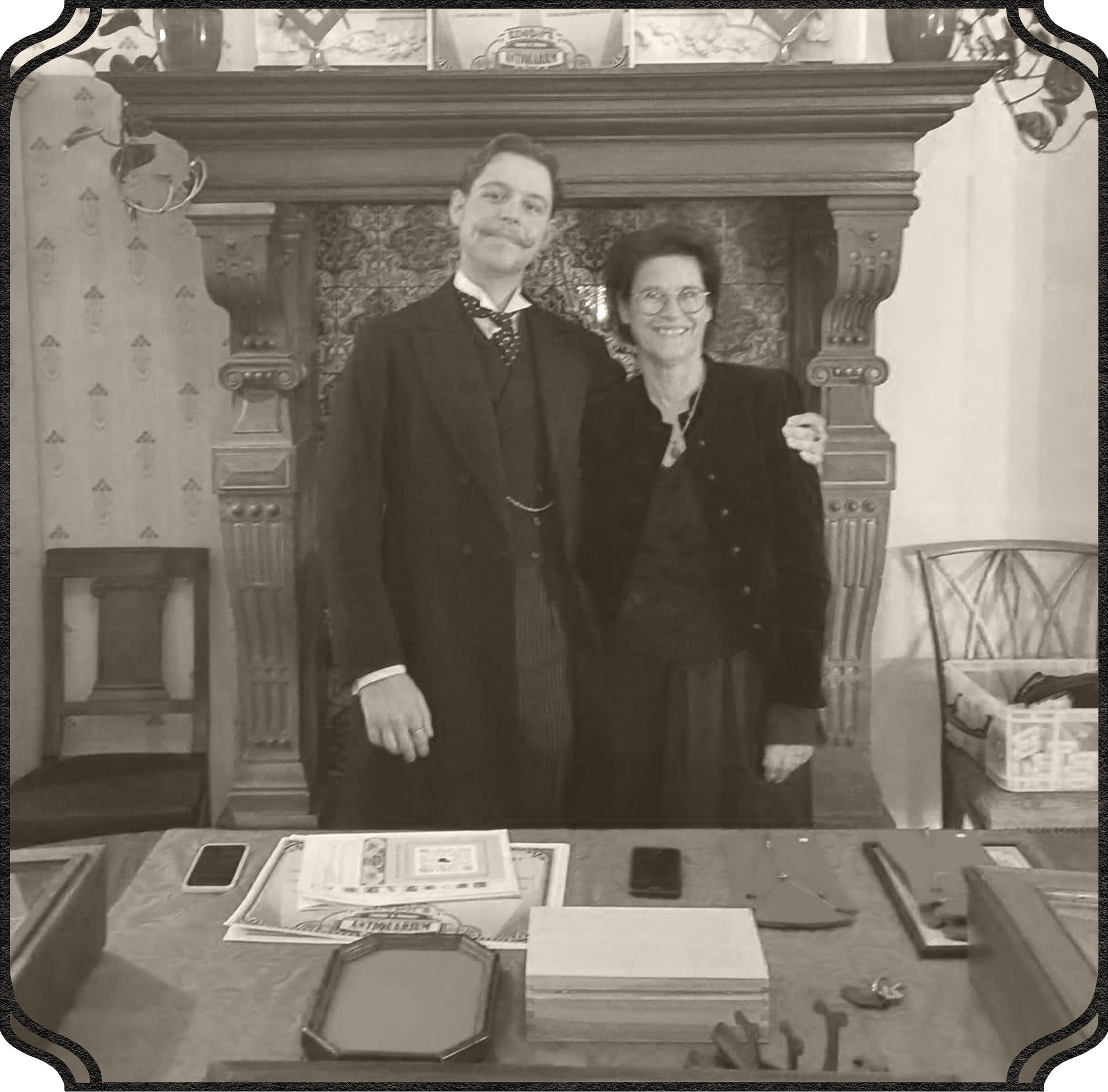 Two people, a man and a woman, smiling and standing behind a display table with various items on it, in front of an ornate fireplace mantel decorated with pictures and ornaments, in a cozy indoor setting.
