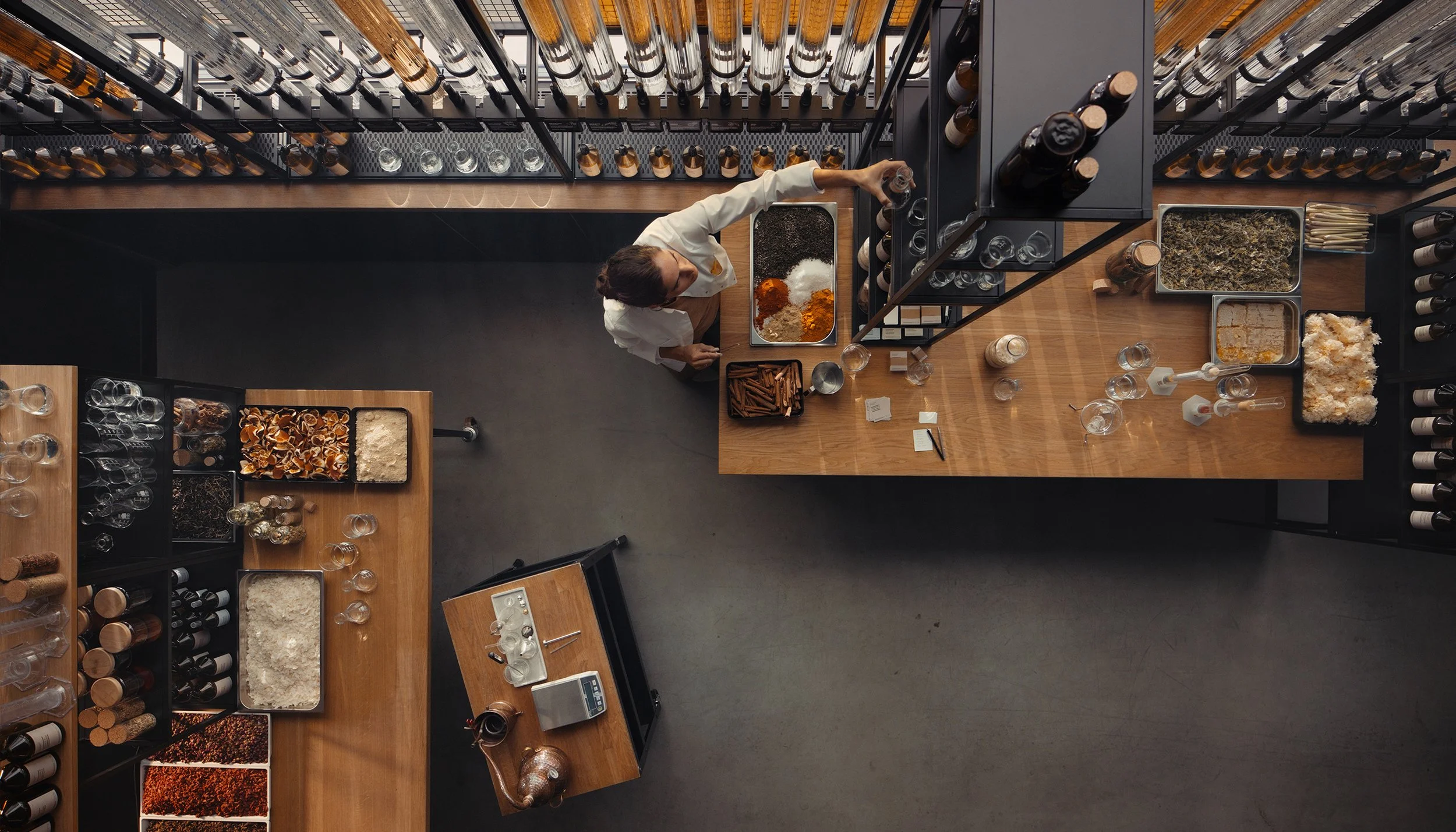 Overhead view of a barista preparing drinks behind a wooden counter at a modern cafe or wine bar, with beverages and snacks on display, and bottles on shelves.