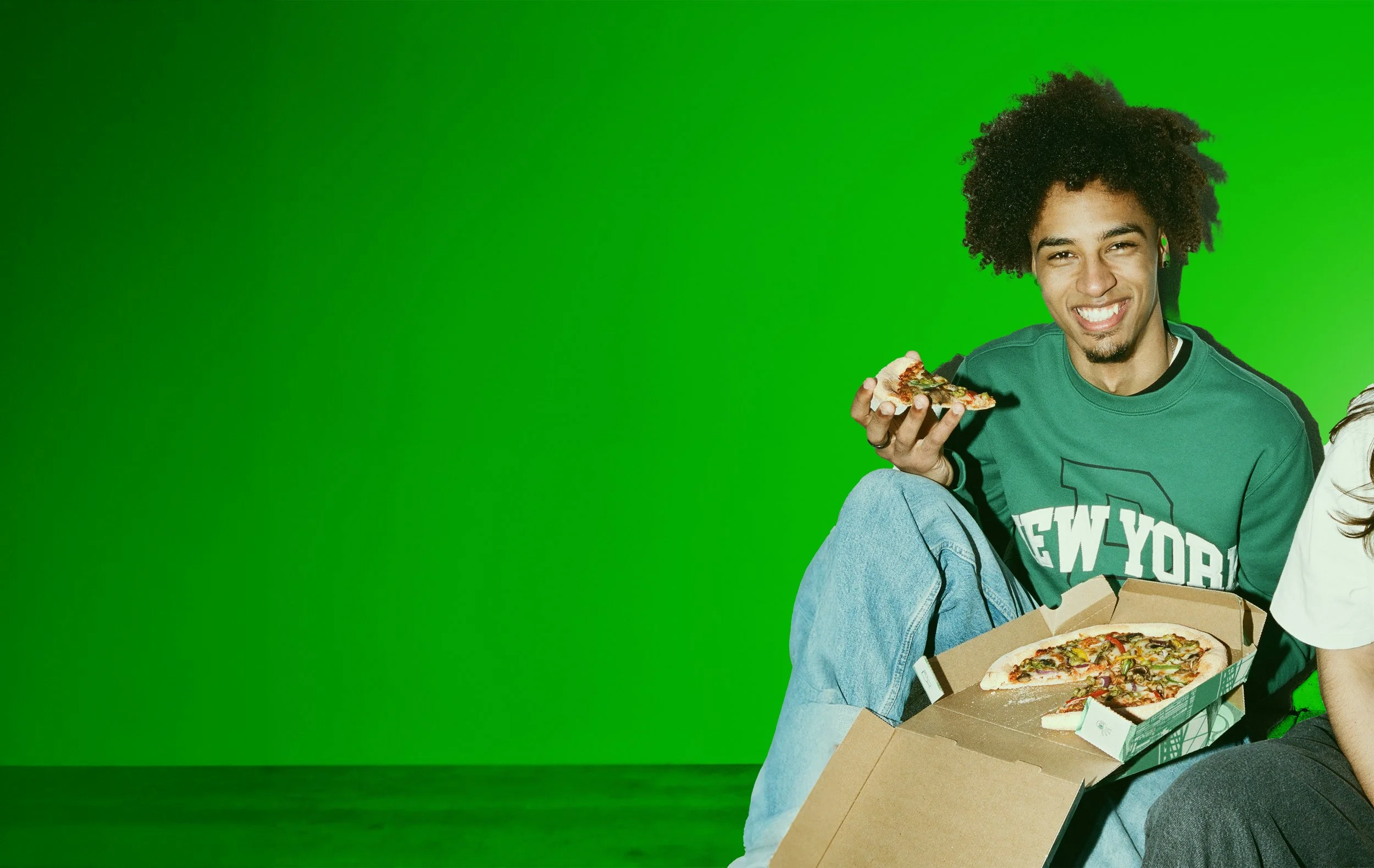 A person with curly hair wearing a green shirt that says 'New York' sitting on the floor with a pizza box and holding a slice of pizza, smiling.