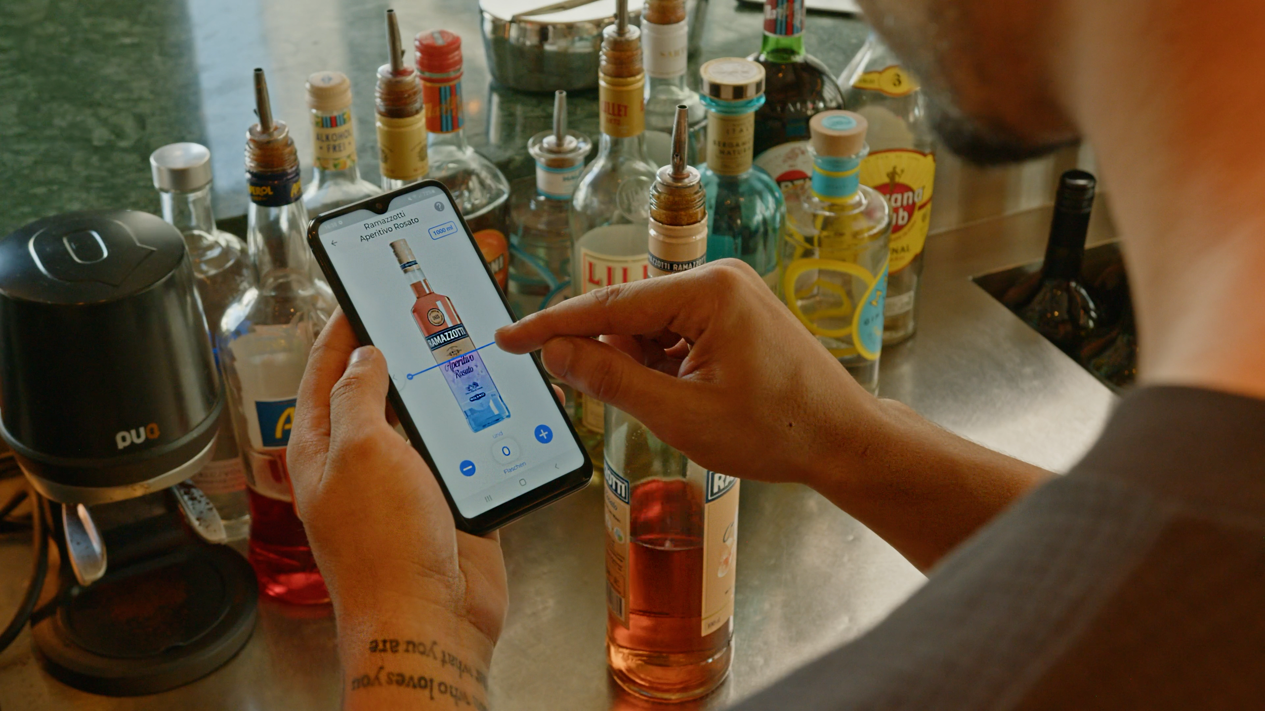 A person is holding a smartphone and using an app to identify a bottle of Ramazzotti Aperitivo Rosato, with various liquor bottles on a bar counter in the background.