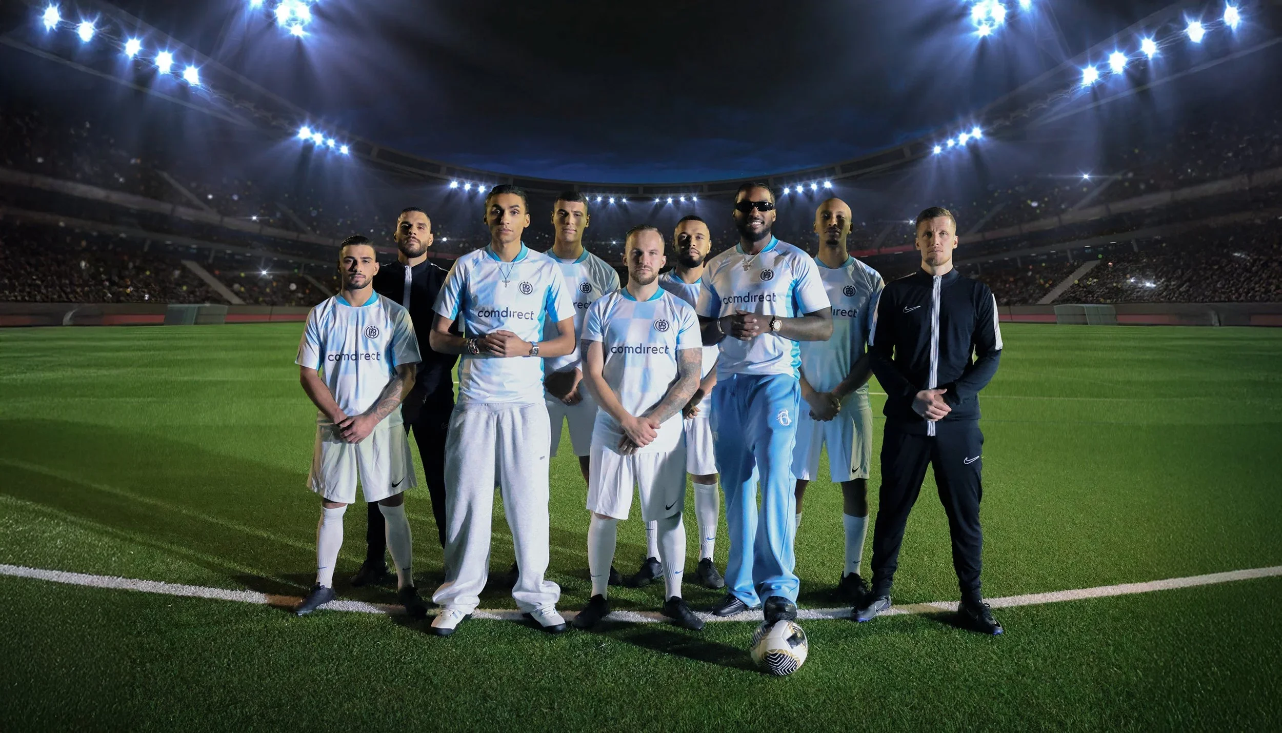Group of soccer players in white and black jerseys standing on a soccer field at night with stadium lights and audience in the background.