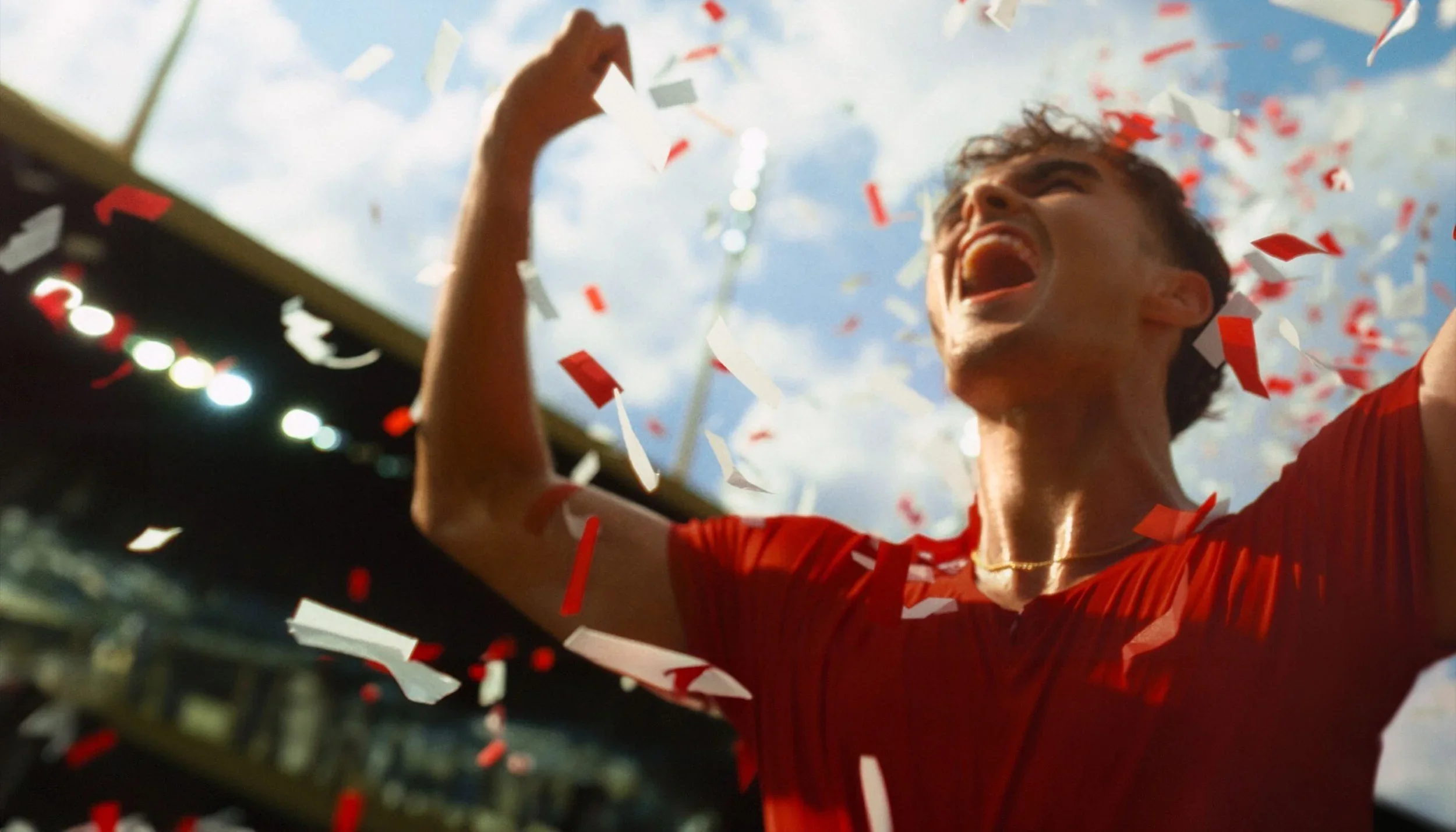 Young man in red shirt celebrating with confetti falling around him at a sports stadium on a sunny day.