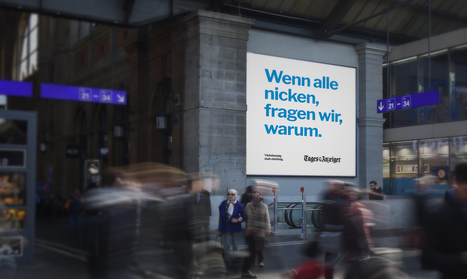 A busy train station with a large illuminated advertisement board displaying the German text 'Wenn alle nicken, fragen wir, warum.' and a subtitle 'Vielstimmig statt einf n'. People are walking through the station, some are blurred due to motion. There are signs indicating platforms 21-34.