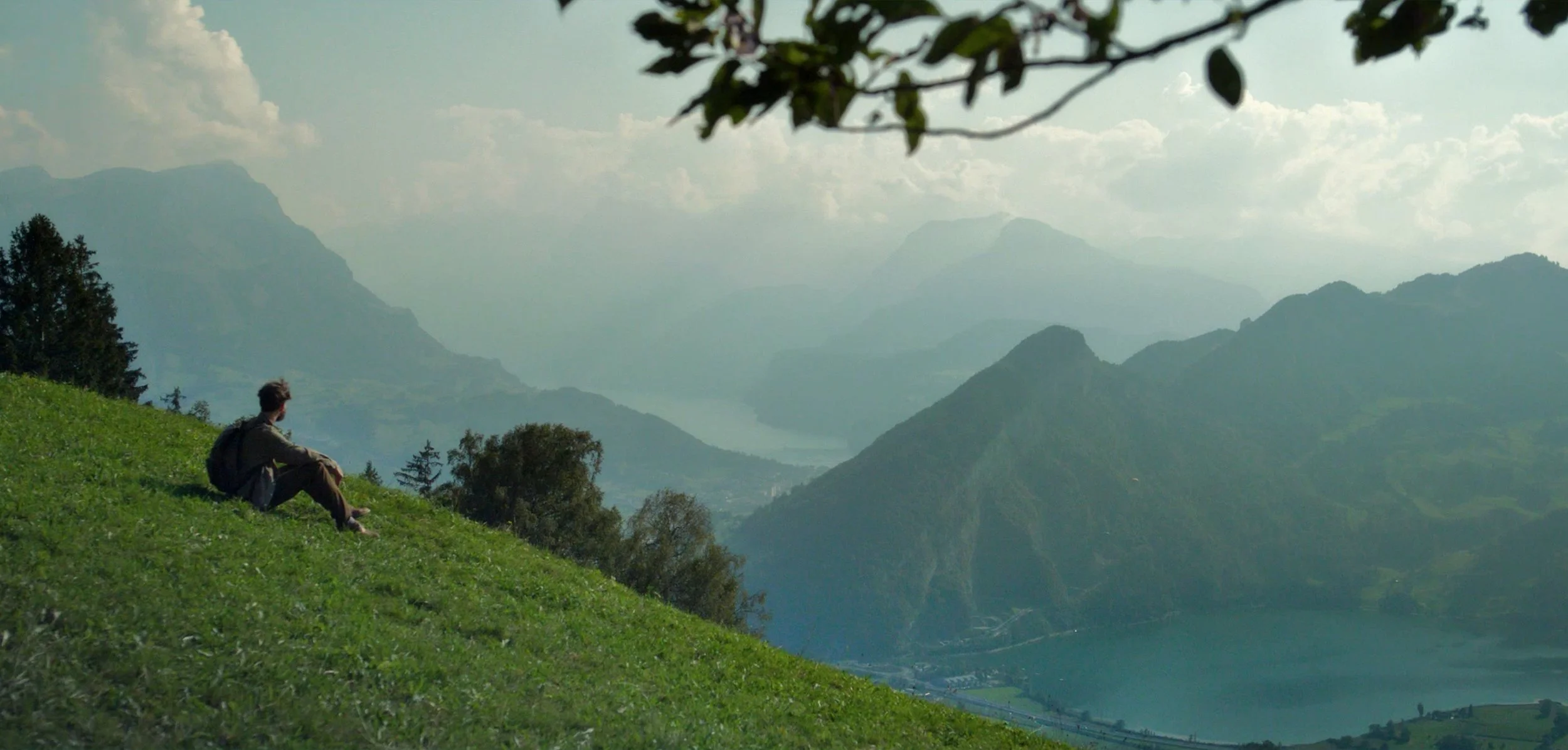 A person sitting on a grassy hillside overlooking a river and mountain landscape with trees and cloudy sky.