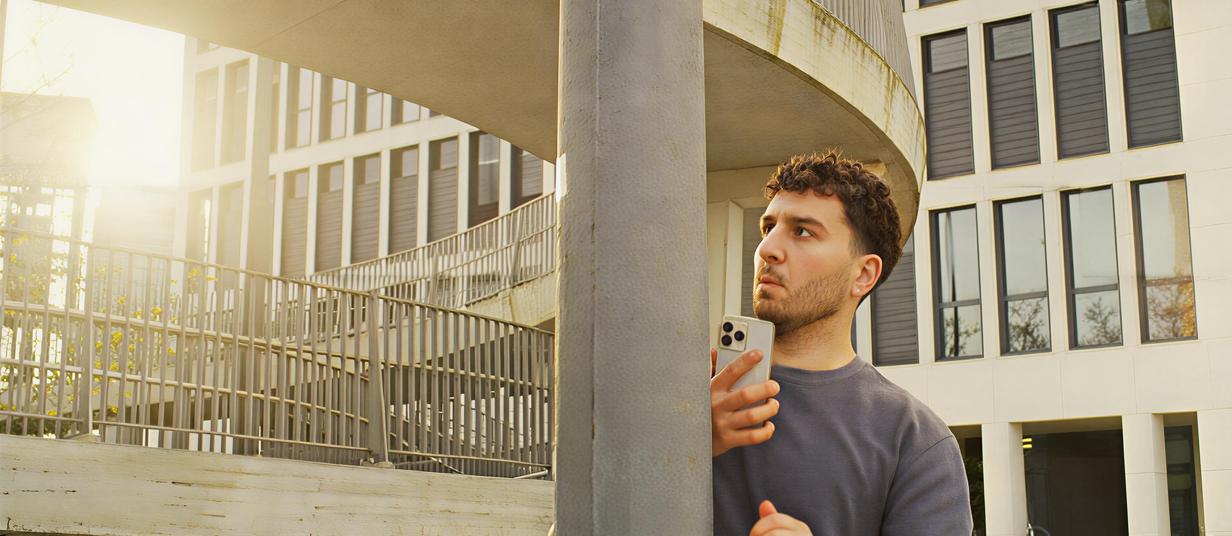 Young man with curly hair and beard holds a smartphone while peering around a concrete column on a city sidewalk, with modern office buildings and a sunny sky in the background.