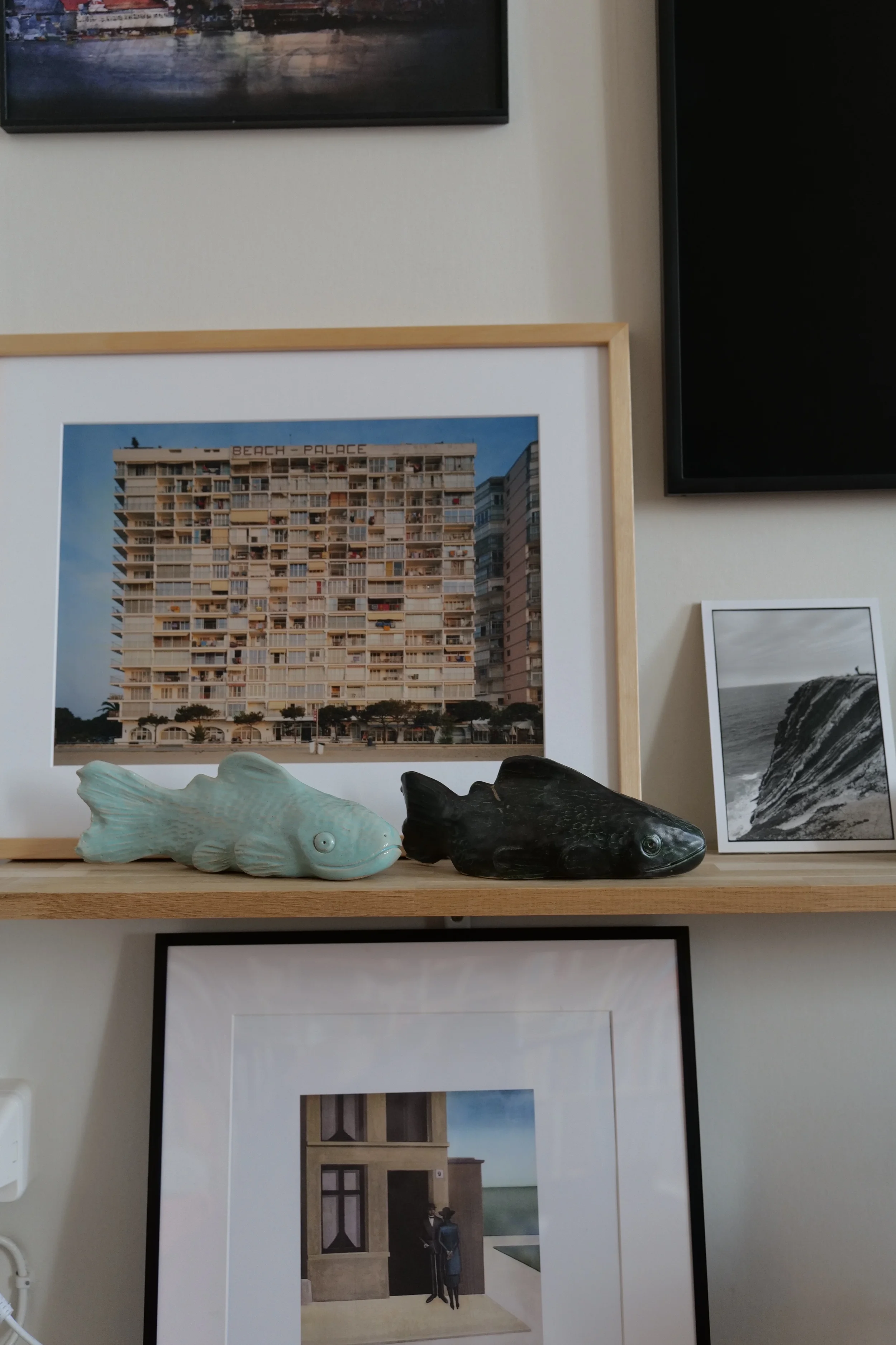 Decorative shelf with two fish sculptures, one light green and one dark black, and framed photographs including a tall building labeled 'Beach Palace', a black-and-white ocean scene, and a painting of a building entrance with two figures.