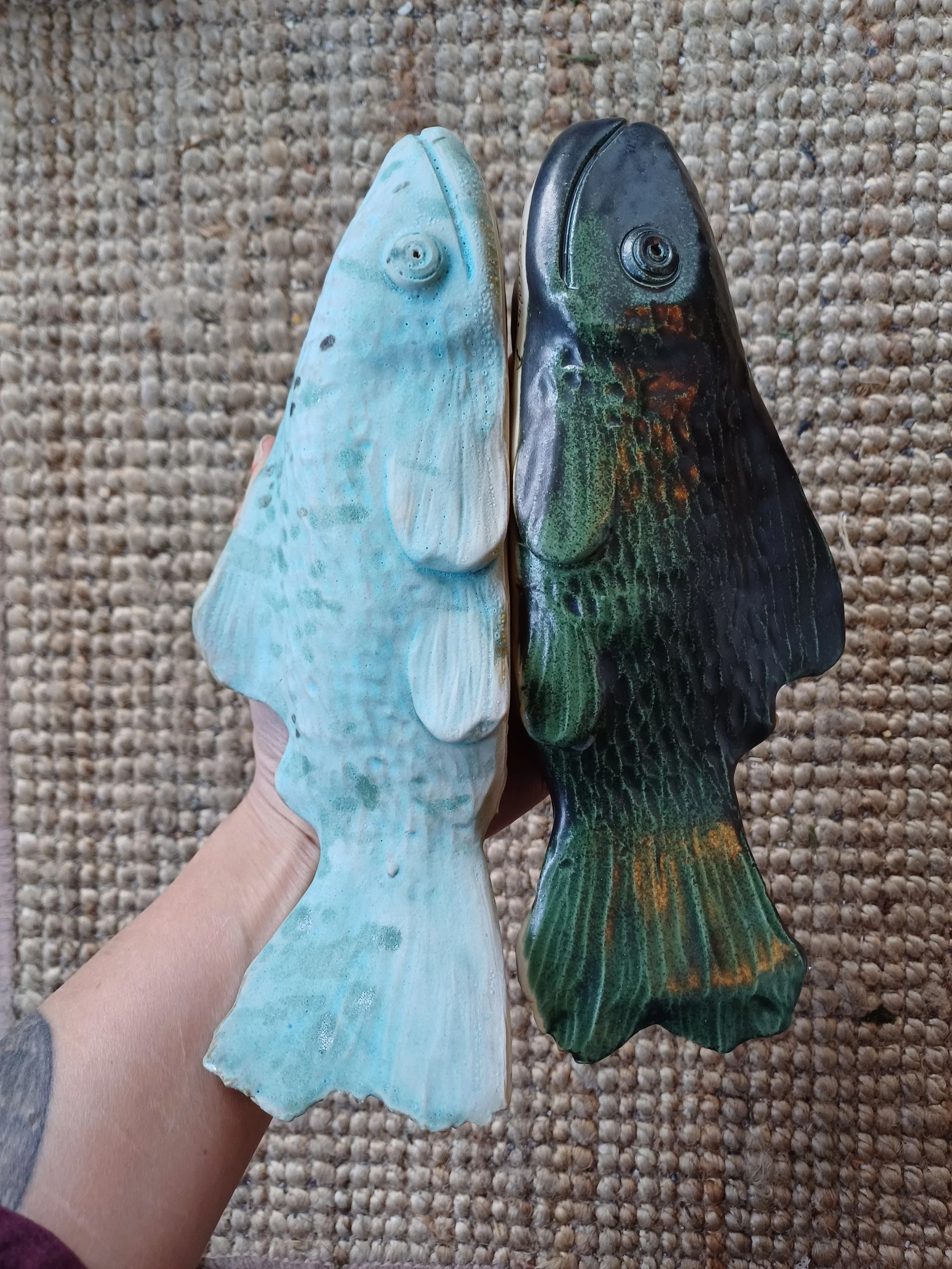Two fish-shaped objects, one light blue and the other dark green with orange, being held in a hand over a textured beige surface.