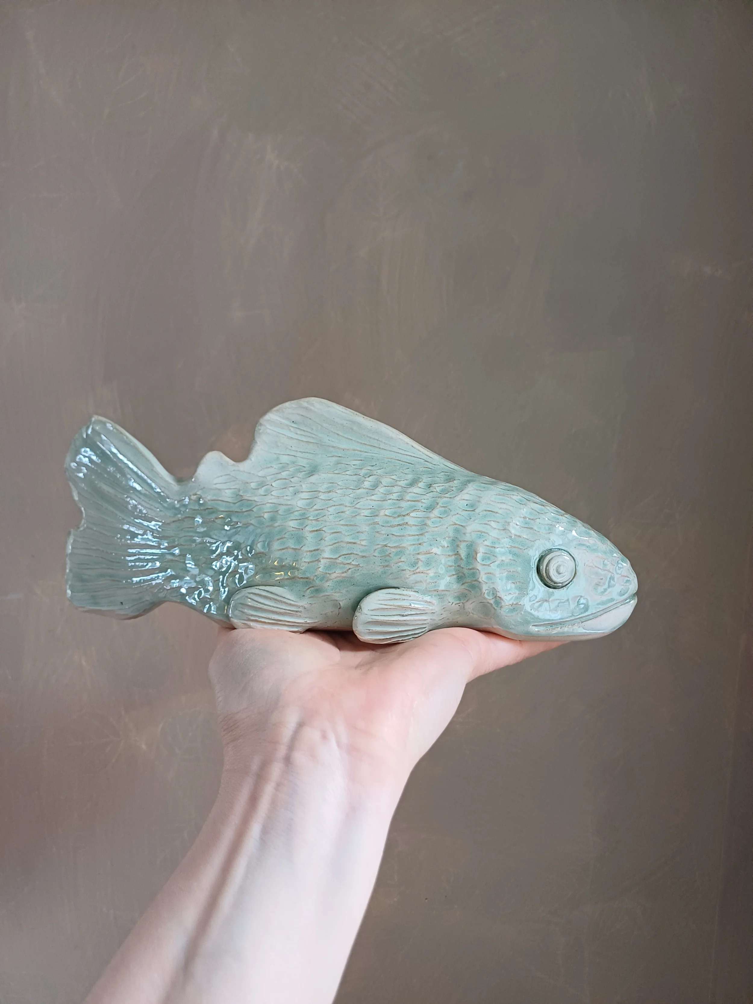 A hand holding a ceramic fish sculpture with a textured, light-colored surface and a spiral detail near its eye, against a plain brown background.