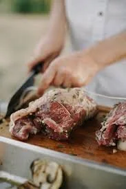 Person slicing raw beef steak on a wooden cutting board.