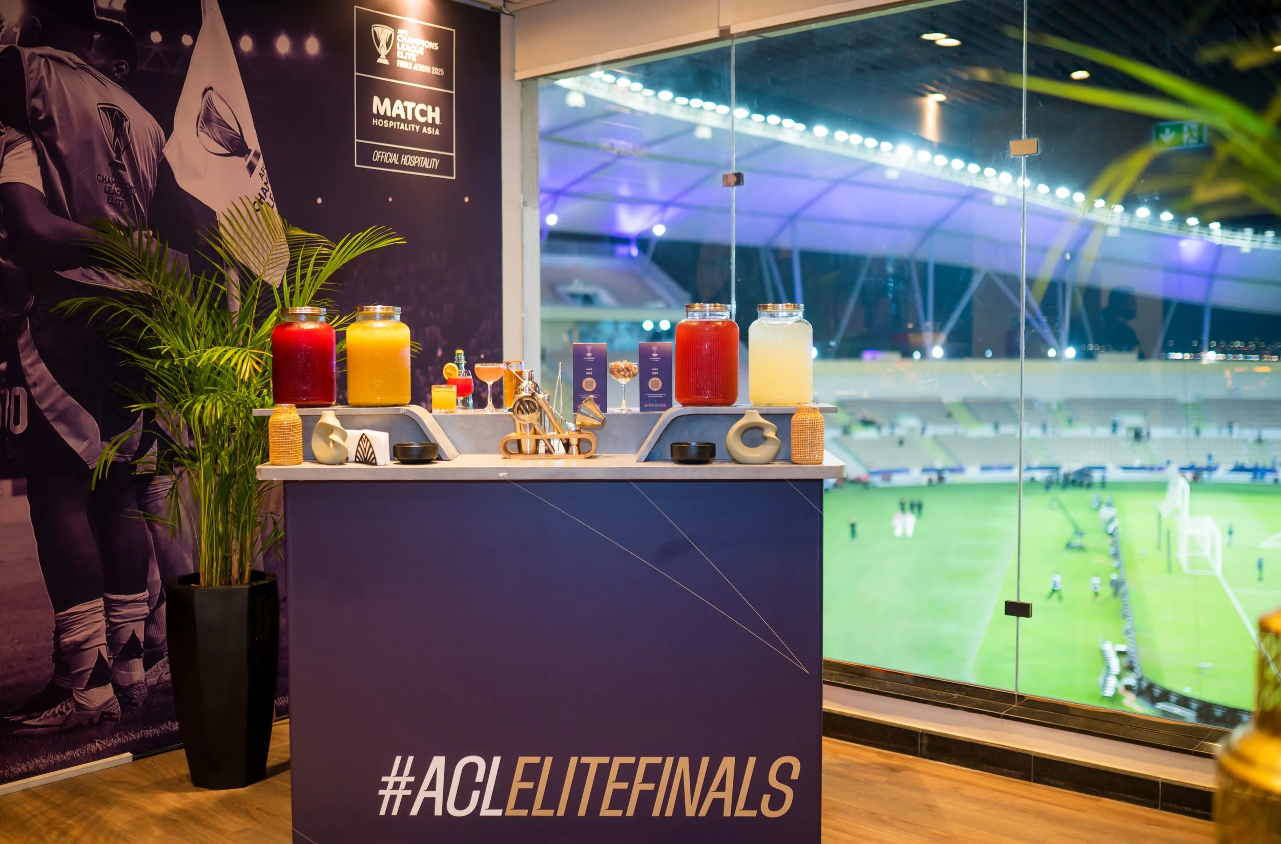 A beverage station with large jars of colourful mocktails, a potted plant, and promotional signage for the AFC Champions League Elite Finals inside Official Hospitality Space.