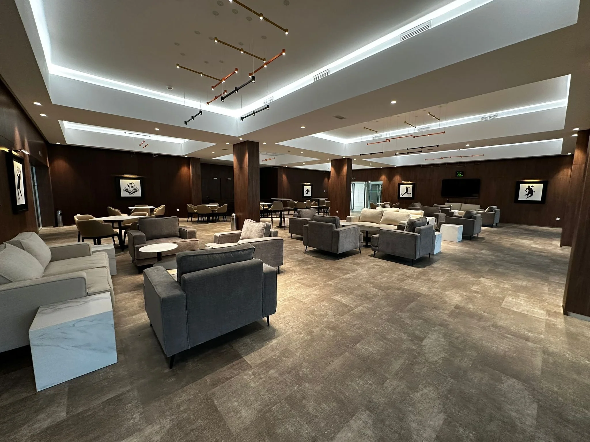 A spacious hotel lounge with modern furniture including gray and beige sofas, armchairs, and small round tables. The ceiling has decorative lighting fixtures resembling linear pendant lights, and artwork depicting sports scenes on the dark wooden walls. The floor is carpeted in a neutral tone, and there are large windows and glass doors in the background.