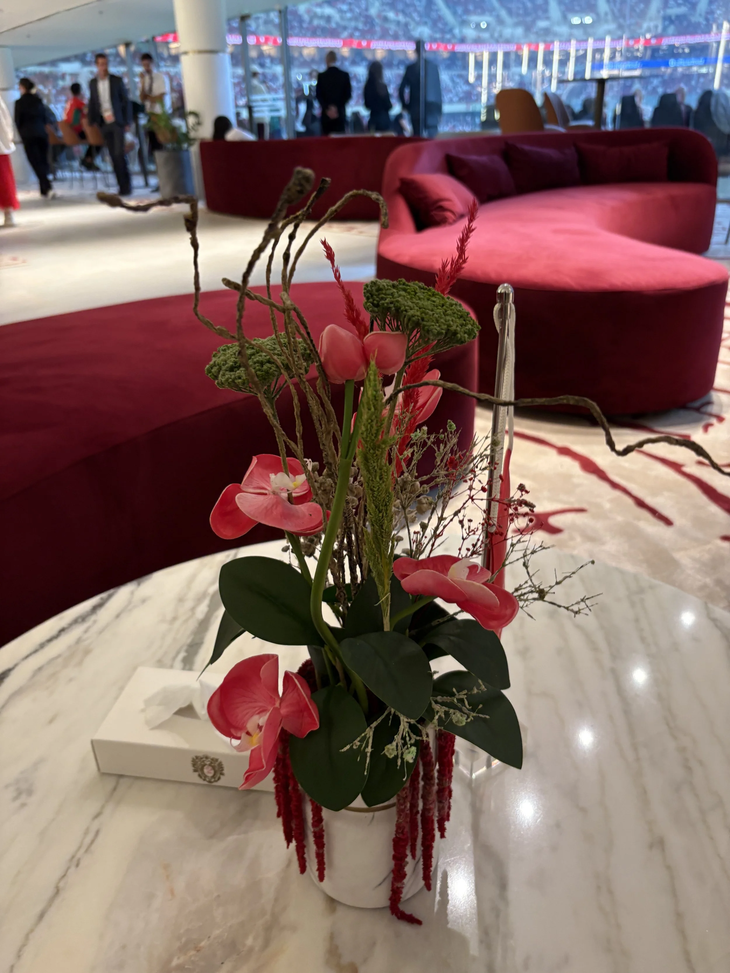 A floral arrangement with pink orchids, green foliage, and twigs in a white vase on a marble table, with a red velvet sofa and a lounge area in the background.
