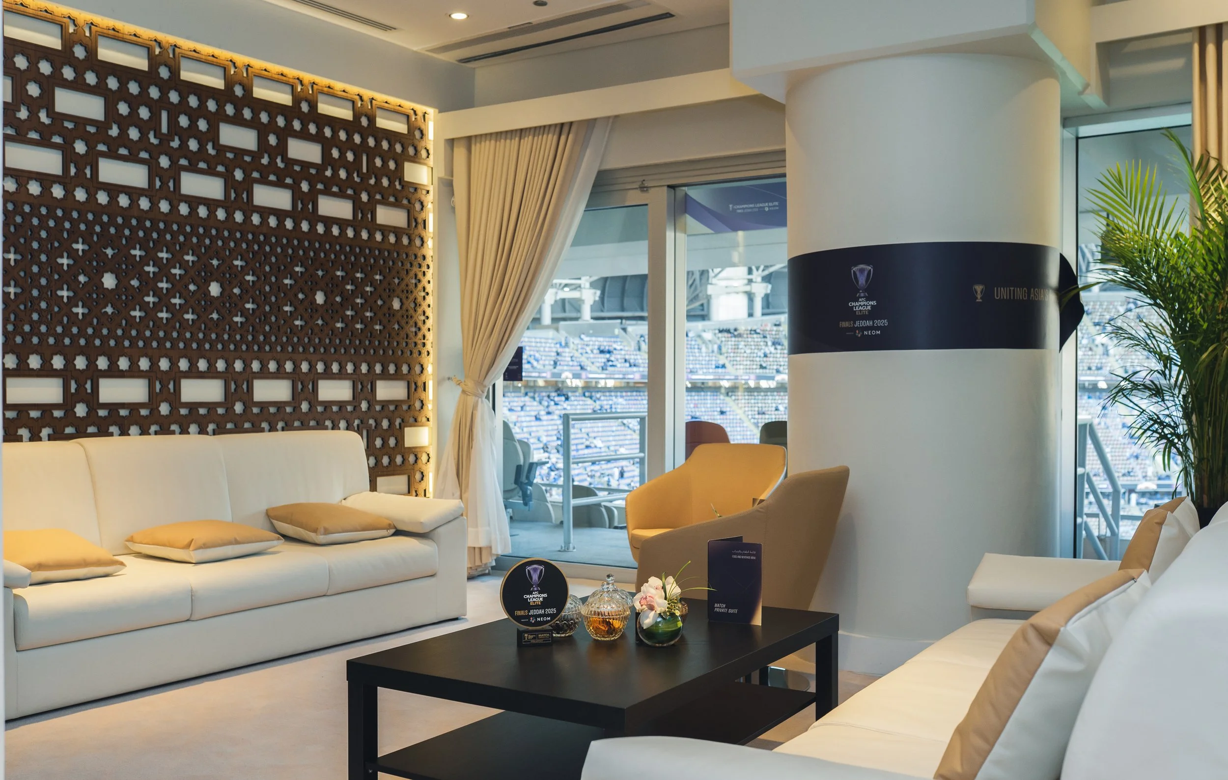 Interior of a luxury Private Suite overlooking a football stadium, with white sofas, a black coffee table holding awards and flowers, and large windows with curtains.