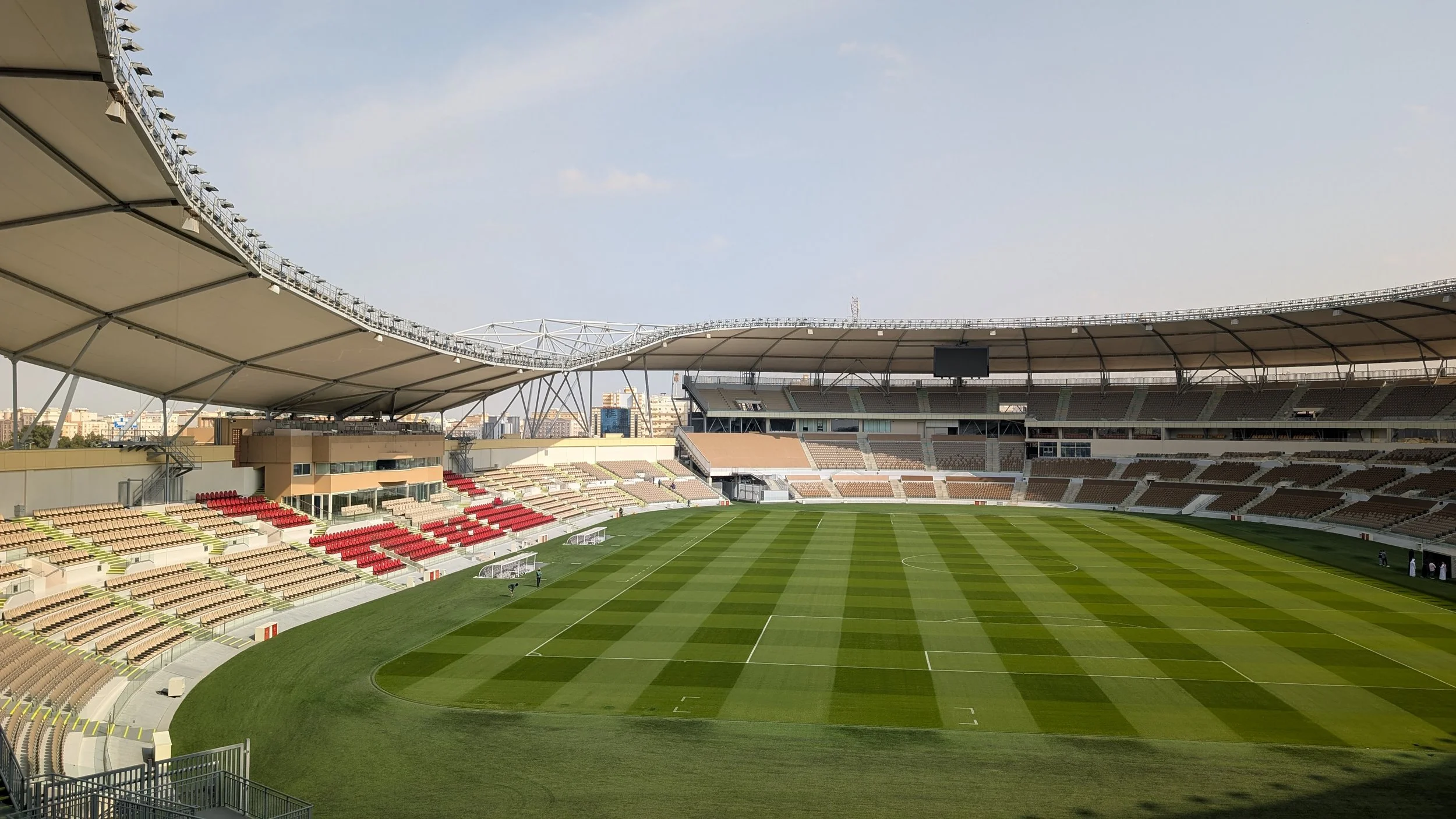 Empty Prince Abdullah Al Faisal Sports City Stadium with a green grass field, surrounded by seating areas and a partially covered roof.