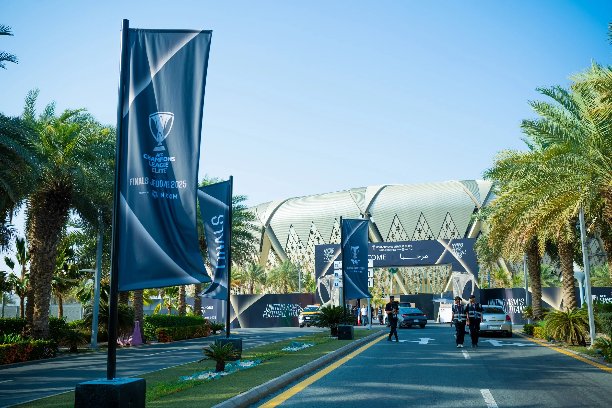 Entrance to the AFC Champions League Elite Finals Jeddah Stadium with flags and signage, surrounded by palm trees, greenery, and a modern building in the background.
