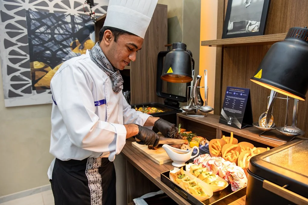 Chef in white uniform and toque preparing food at private suite buffet station with various dishes and decorative elements.