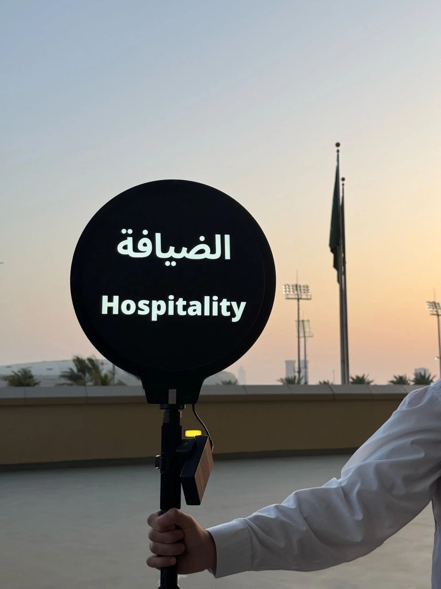 A person holding a black sign with white text that reads 'Hospitality' in English and Arabic script, against a backdrop of a sunset sky, flag, and stadium lights.