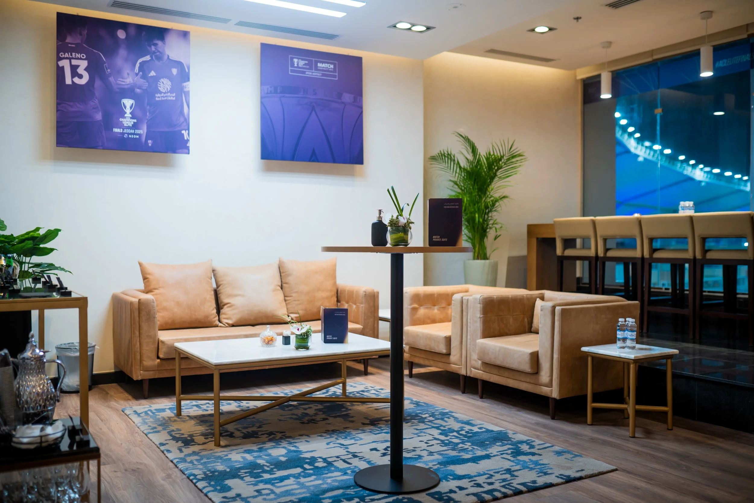 A cosy private suite area with beige sofas, a white coffee table, green plants, and wall décor, near a high table with high chairs and a large window overlooking the stadium.