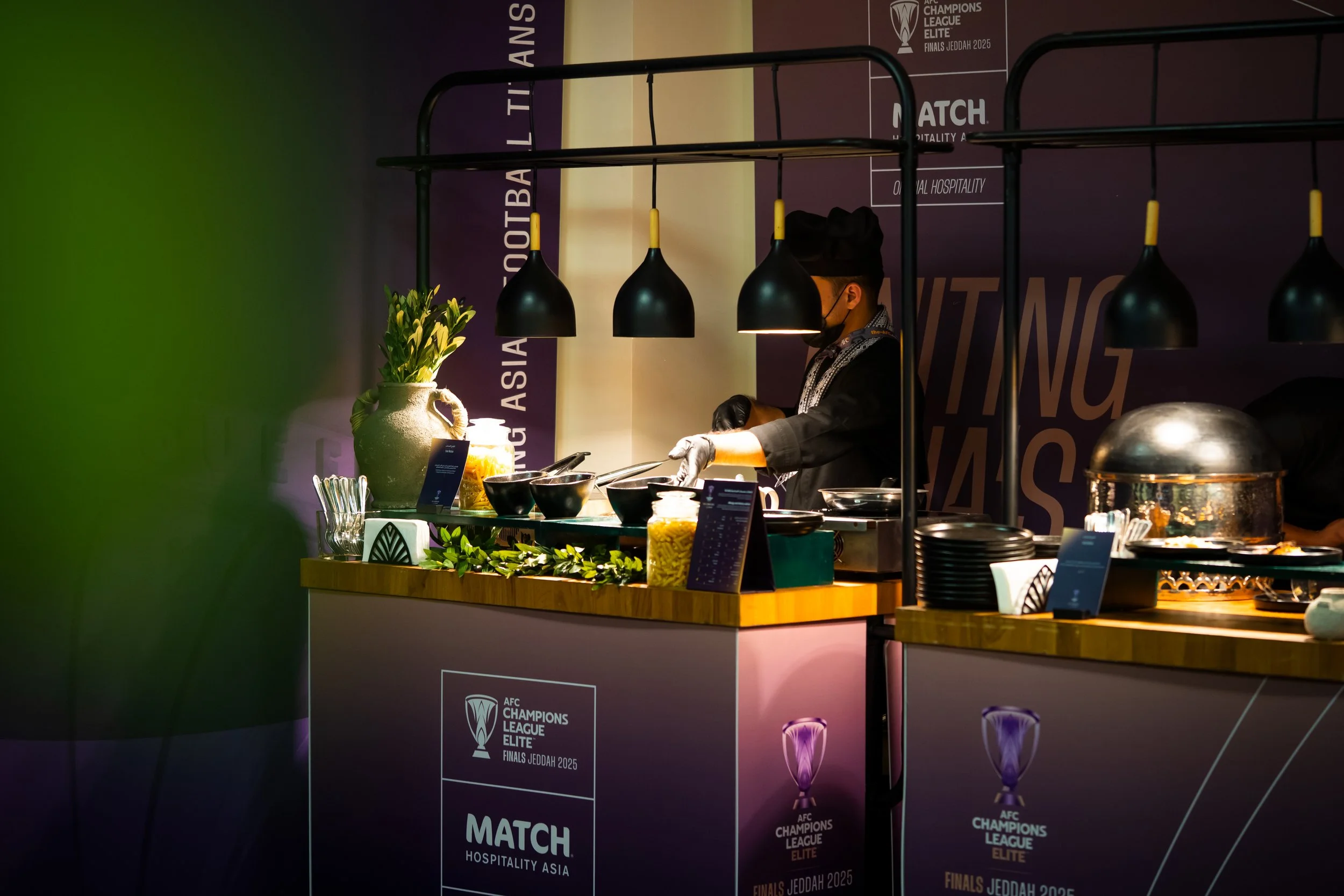A chef preparing food at a buffet station with black pendant lights, a vase with flowers, and a backdrop with AFC Champions League Elite Finals Jeddah branding.