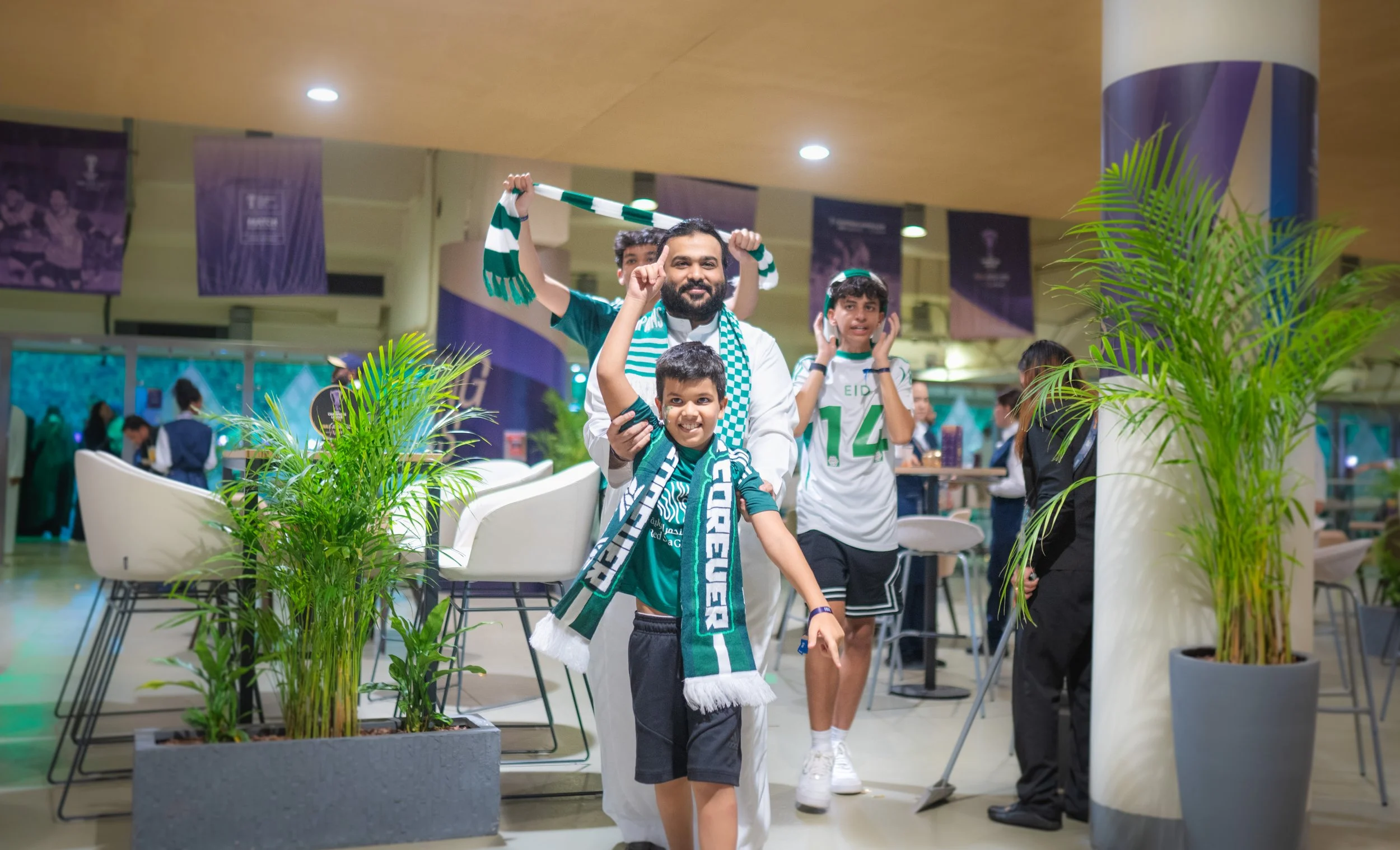 A group of people, including children and an adult, wearing green and white sports fan apparel, gathering in an indoor venue decorated with banners, plants, and seating areas. The group appears excited and happy, celebrating football with scarves and gestures.