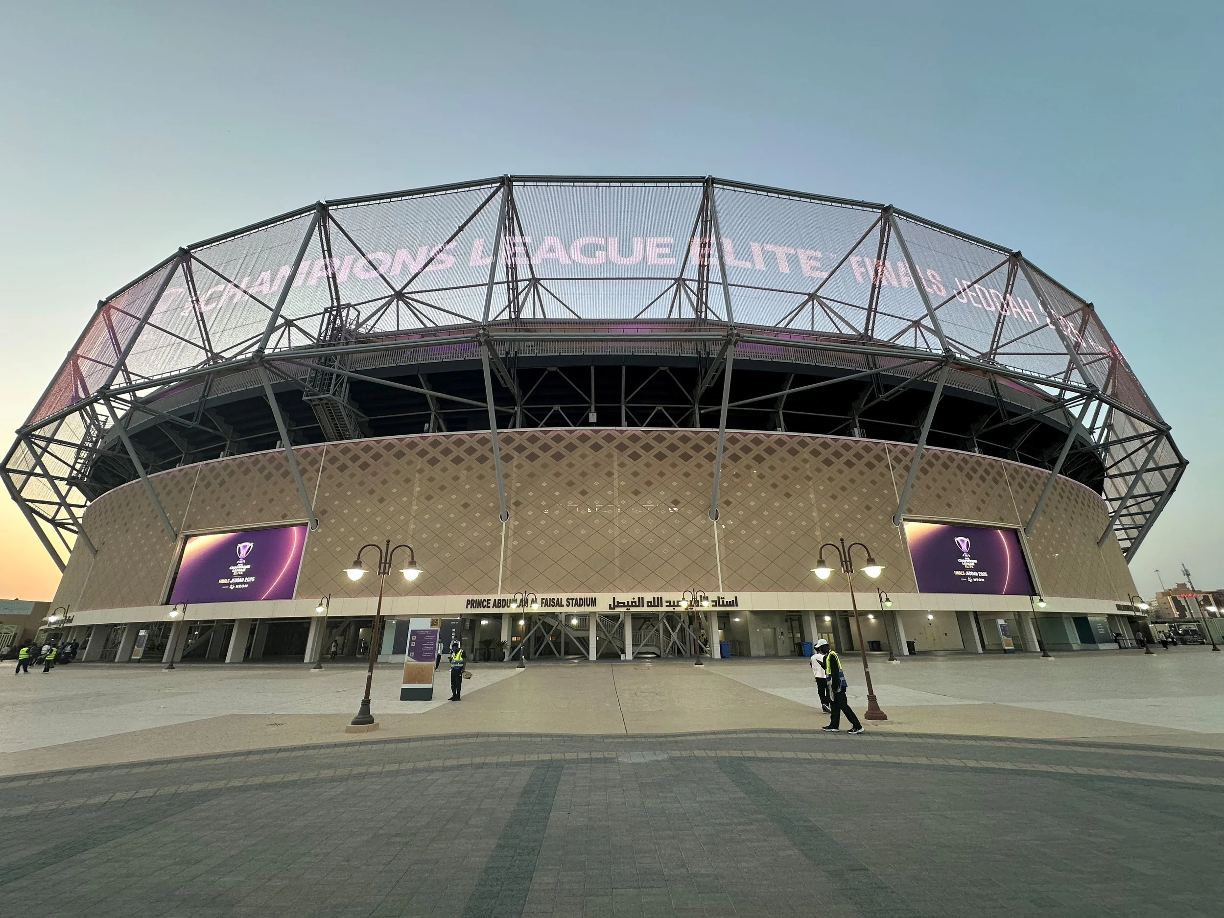 Exterior view of Prince Abdulaziz Faisal Stadium with large electronic banners displaying AFC Champions League Elite Finals Jeddah and football match information in Arabic, at dusk.
