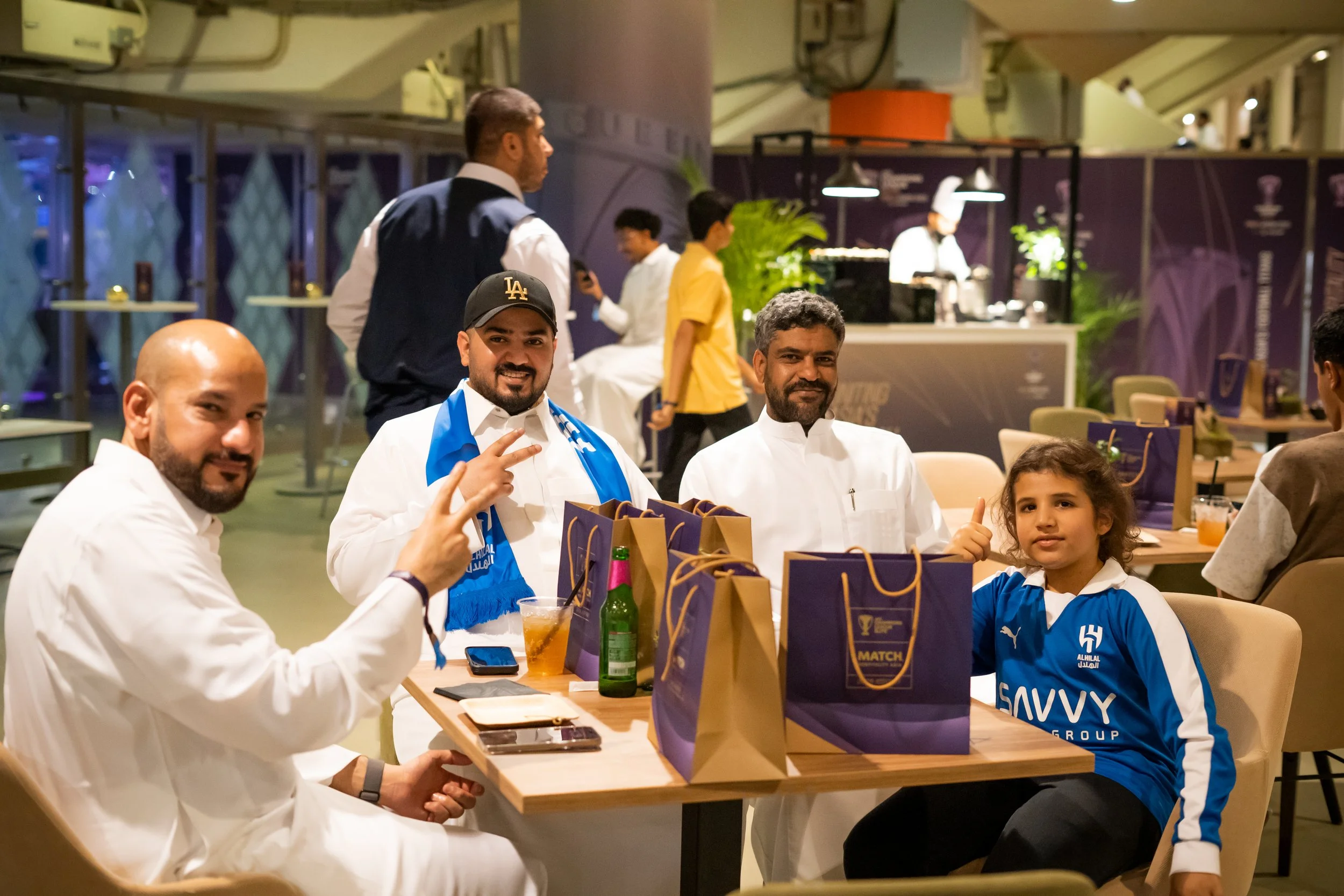 Official Hospitality guests sitting at a table in MATCH Club Gold, with gift bags on the table, a drink, and a phone, with a chef in the background.