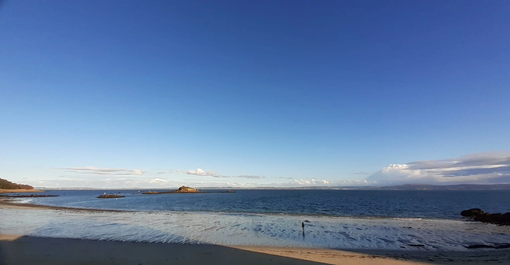 Plage avec sable, mer calme, quelques rochers, ciel bleu avec quelques nuages, horizon maritime, personne marchant sur la plage