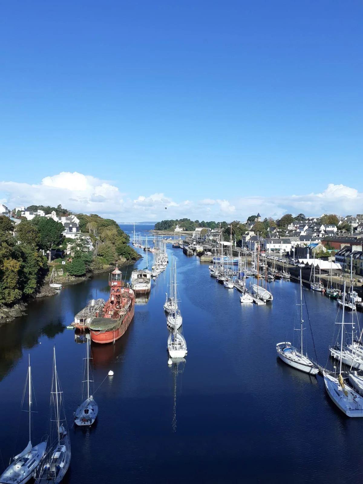 Port de plaisance avec de nombreux bateaux et voiliers, surmonté d'un ciel bleu avec quelques nuages, entouré de maisons résidentielles.
