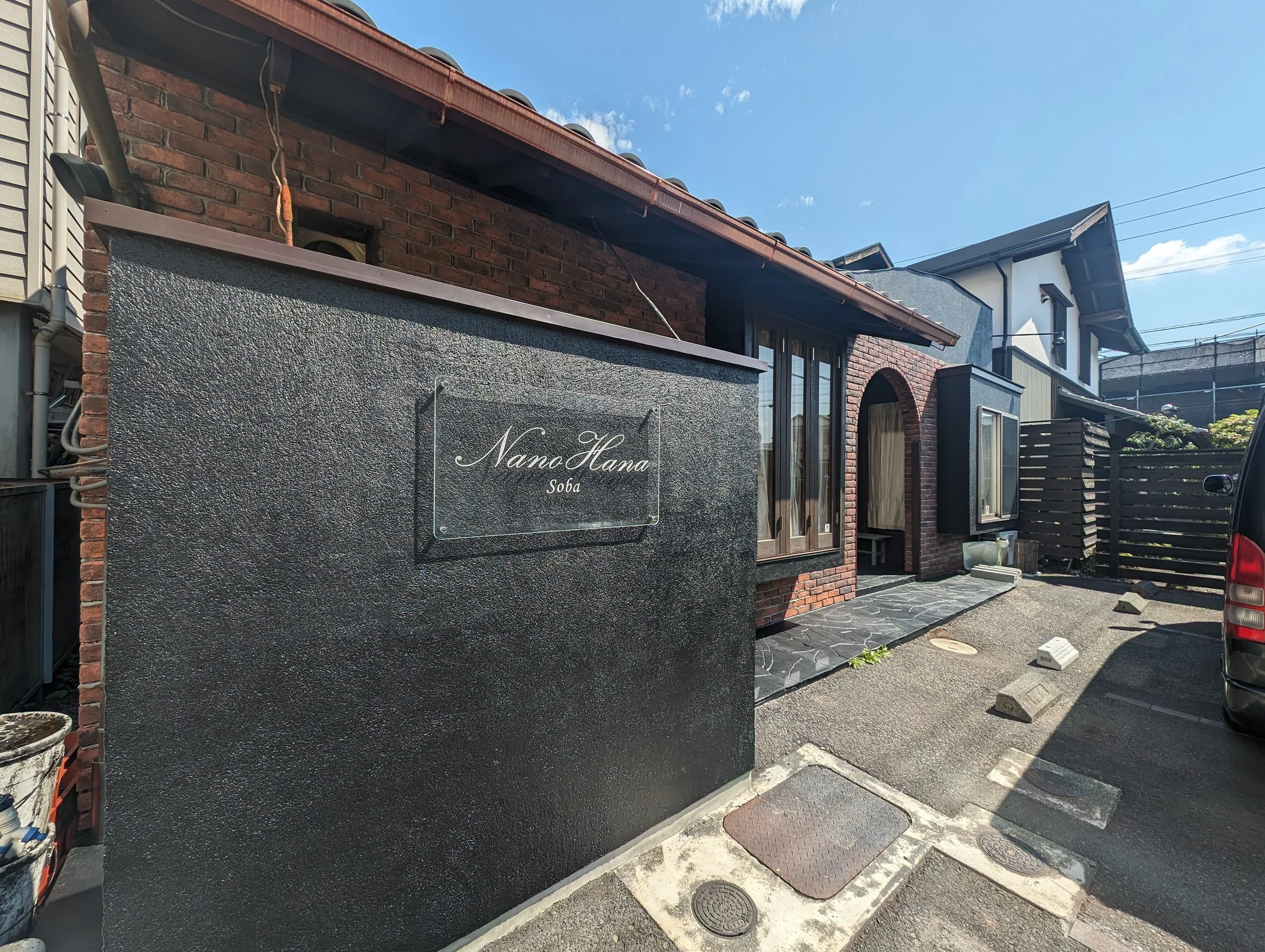 黒い看板に白い文字で「Nano Hana Soba」と書かれているレンガ造りの建物の正面。青空と一部の建物が見える。