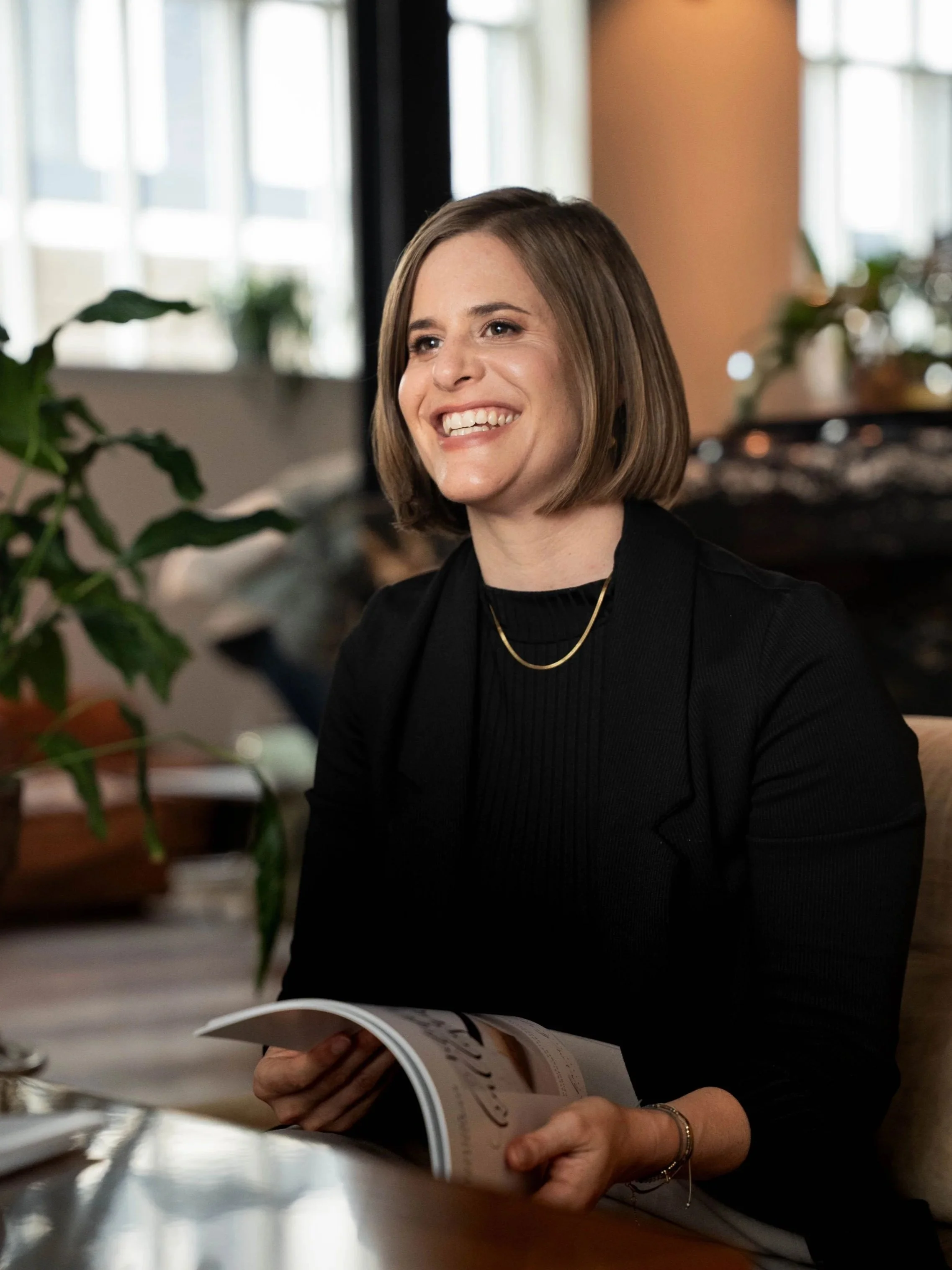 Eine lächelnde Frau mit Kurzhaarfrisur, schwarze Kleidung und Goldschmuck, sitzt in einem Raum mit großen Fenstern, hält eine Zeitschrift in den Händen und scheint im Gespräch zu sein.