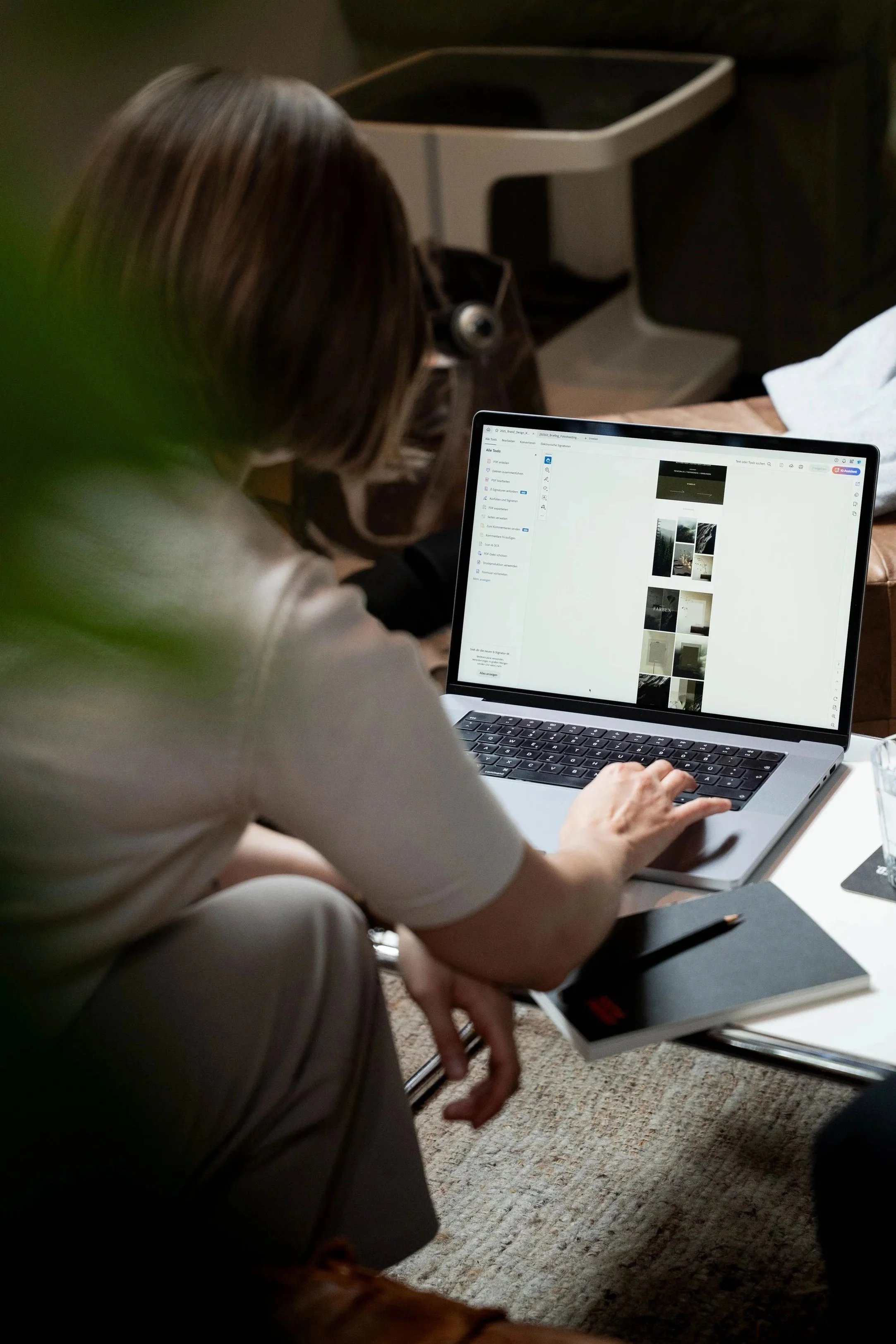 Eine Frau sitzt an einem Tisch und benutzt einen Laptop, auf dem sie eine Fotosammlung betrachtet. Ein Notizbuch mit Stift liegt vor ihr auf dem Tisch.