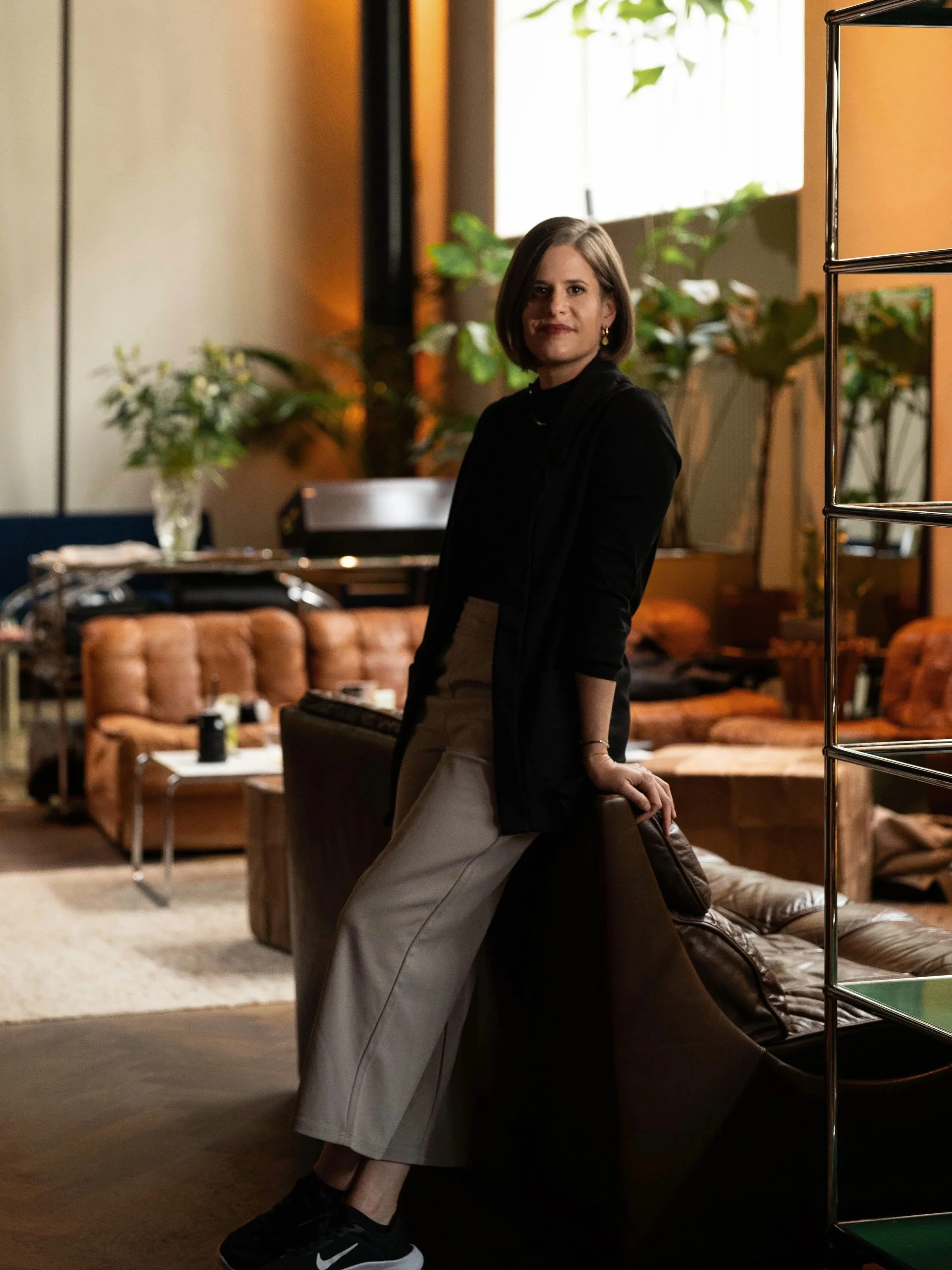 Eine Frau mit braunen Haaren trägt schwarze Kleidung und beige Hosen, steht in einem gemütlichen, stilvollen Raum mit Sofas und Pflanzen.
