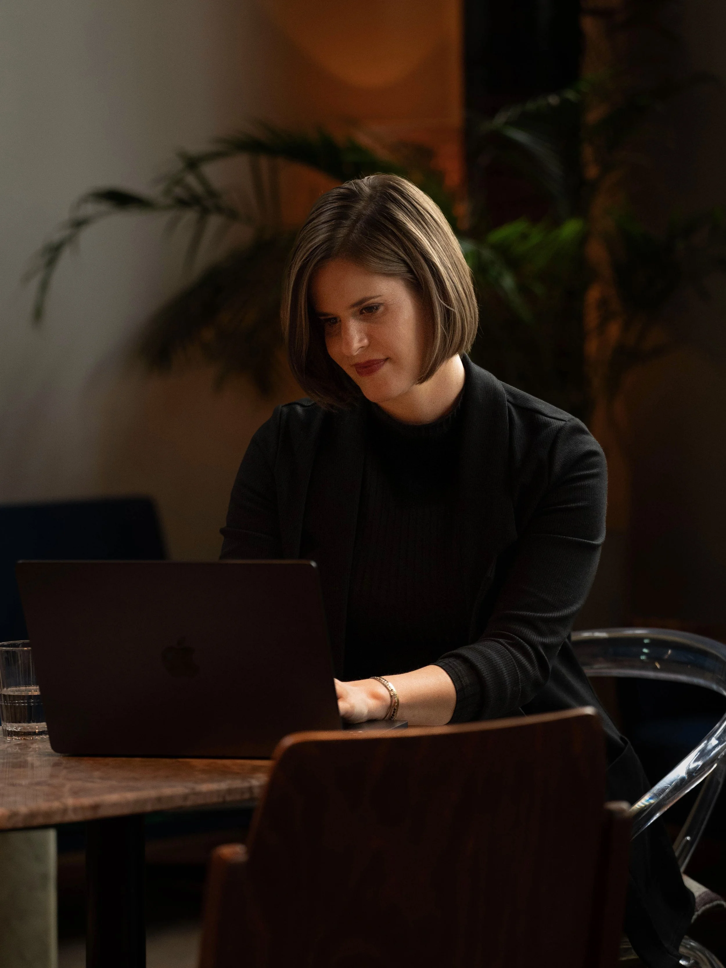 Frau sitzt an einem Tisch und arbeitet an einem schwarzen MacBook in einem Raum mit Pflanzen im Hintergrund.
