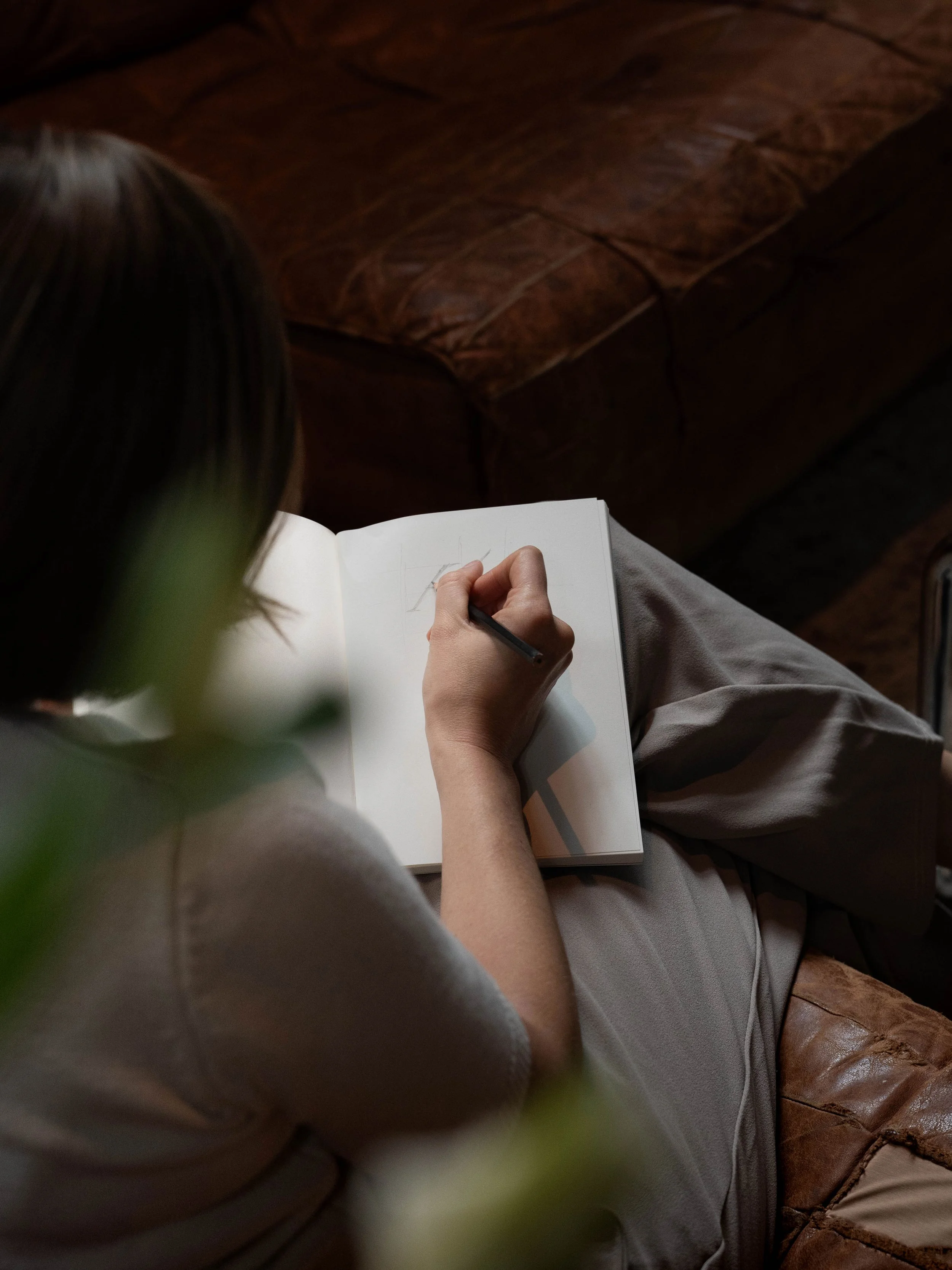 Person schreibt in ein offenes Notizbuch, sitzend auf einem braunen Ledersofa.