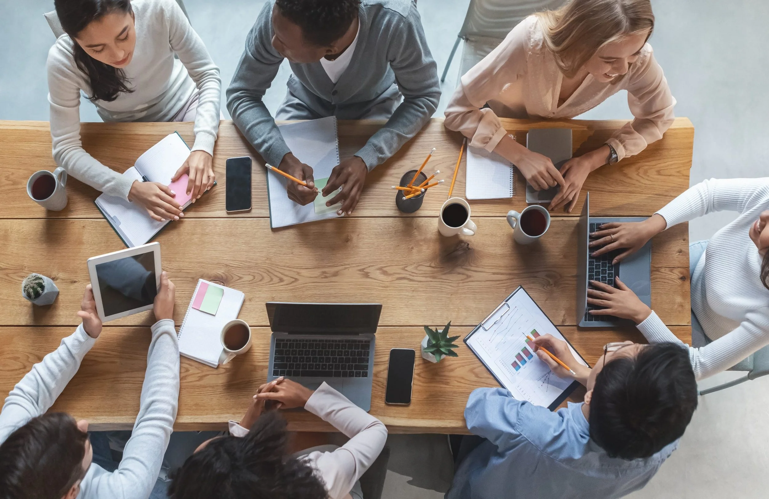 A top-down view of six people gathered around a large wooden table, working on laptops, tablets, and notebooks with coffee cups and stationery, engaged in a collaborative meeting or discussion.