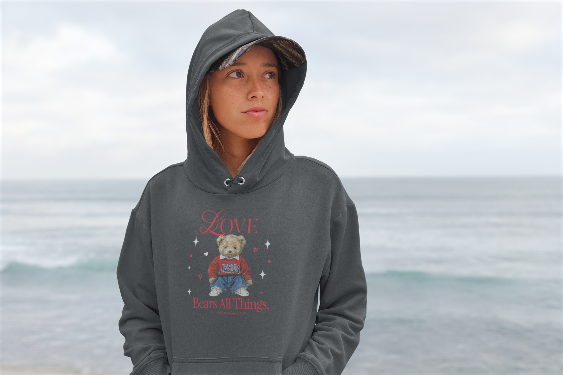 A young woman with a hoodie and camo cap standing on a beach with the ocean in the background. She is wearing a gray hoodie with a graphic of a teddy bear and the words 'Love Bears All Things' on it.