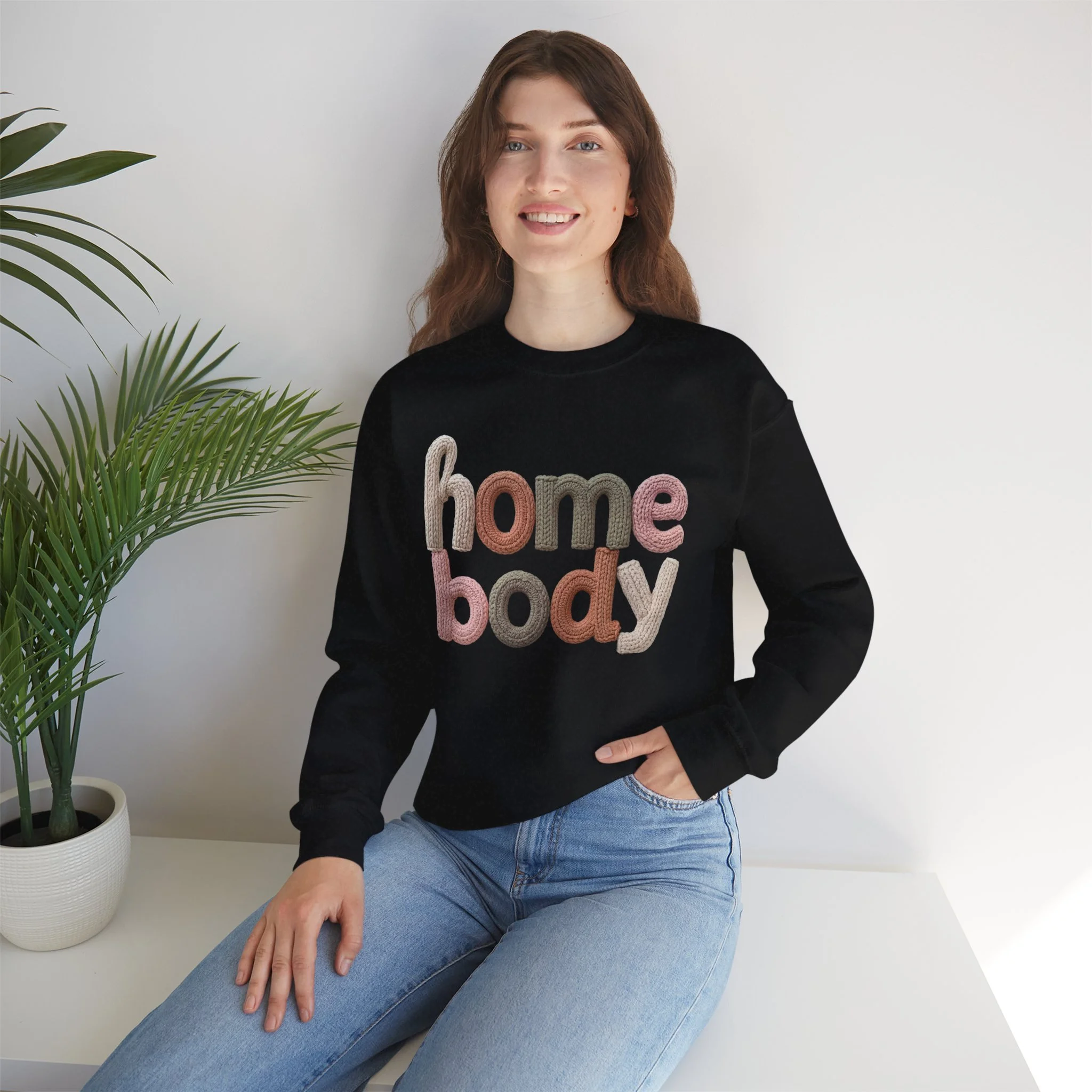 Woman sitting on a white surface next to a potted plant, wearing a black sweatshirt with the words 'home body' in colorful embroidered letters.