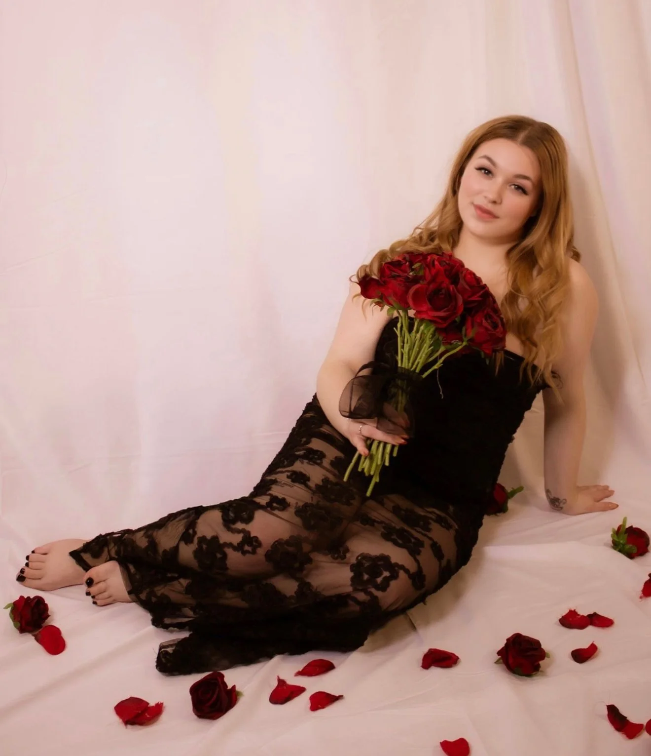 A woman with long, wavy red hair sitting on a white sheet, holding a bouquet of red roses, wearing a black lace dress, surrounded by scattered red rose petals.