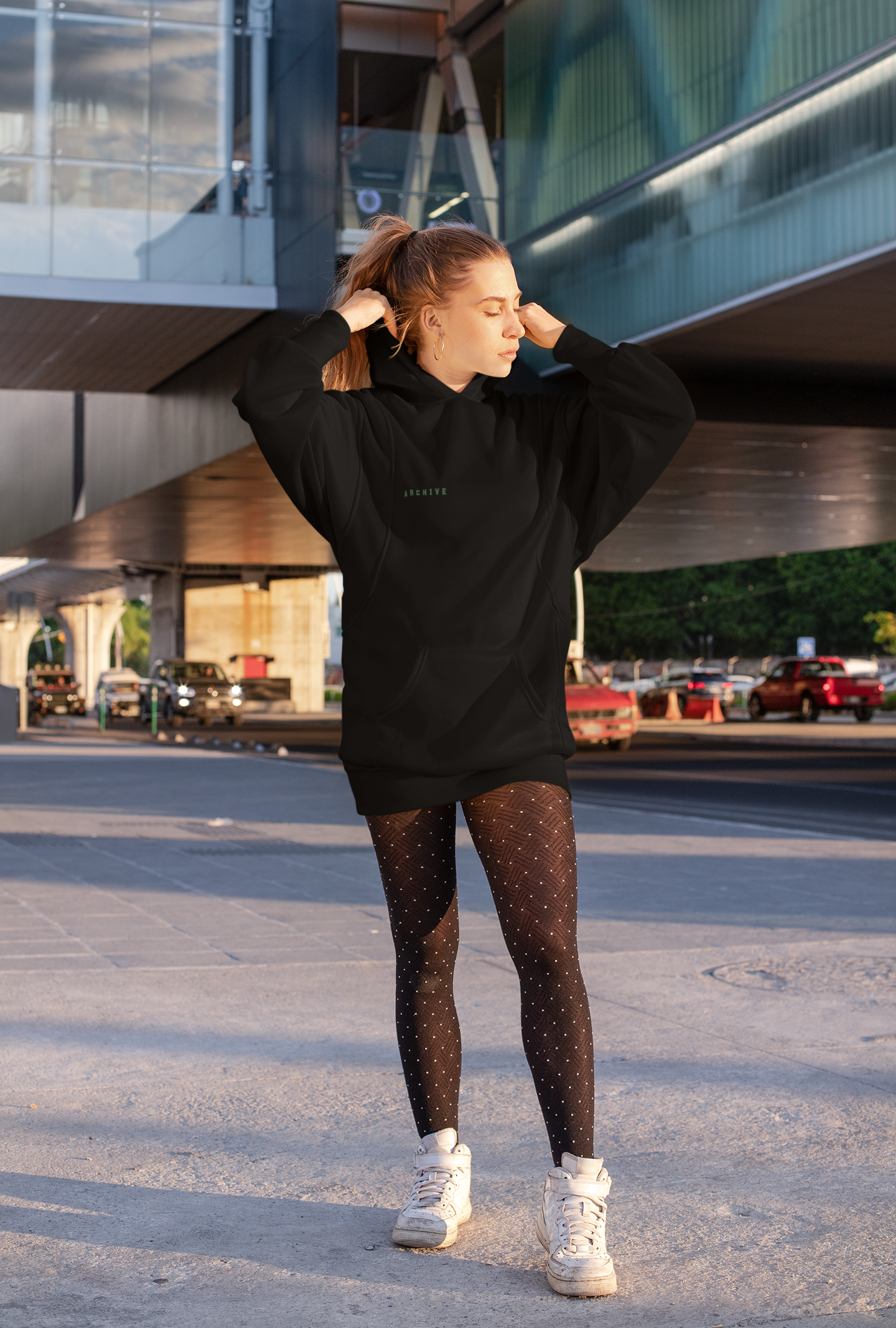 A young woman with red hair in a ponytail, wearing a black hoodie with green text and patterned black tights with white dots, is standing on a city street during sunset, stretching her arms behind her head.
