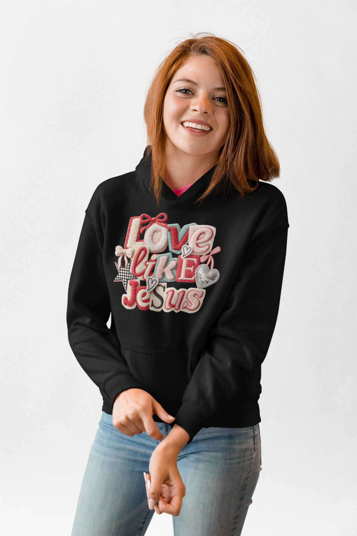 Young woman with red hair smiling, wearing black hoodie with colorful embroidered words and designs, and light blue jeans, standing against a white background.