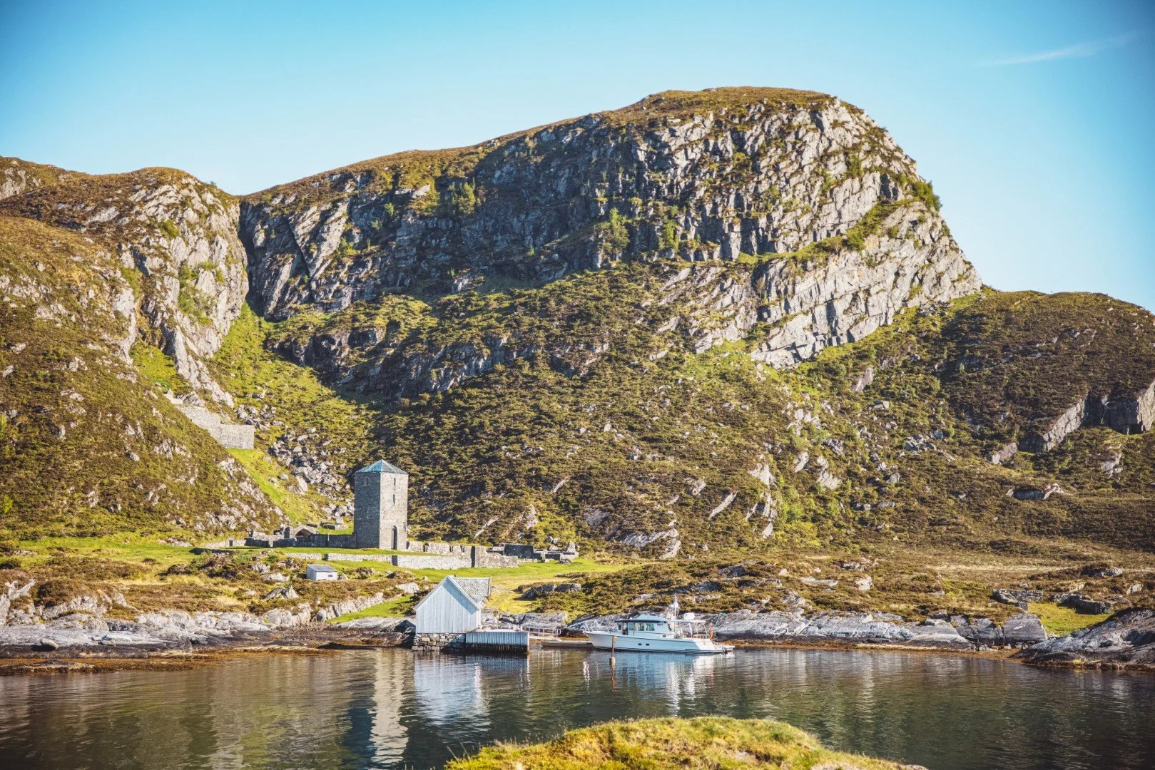 A scenic view of a rocky hillside with a small stone building, a boat dock, and a boat by the water, under a clear blue sky.