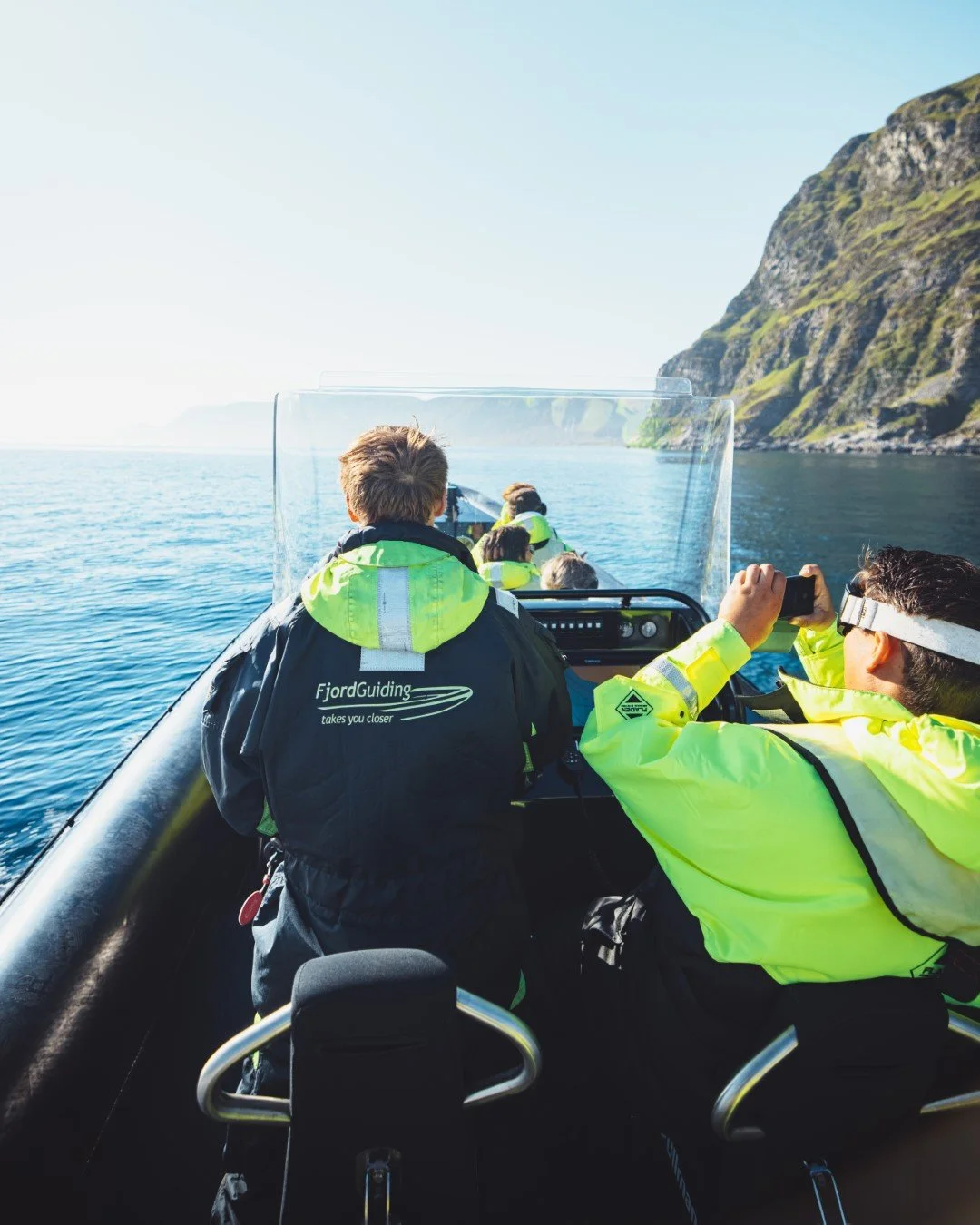 People riding a boat on a body of water with green, rugged cliffs in the background, wearing bright yellow and black jackets.