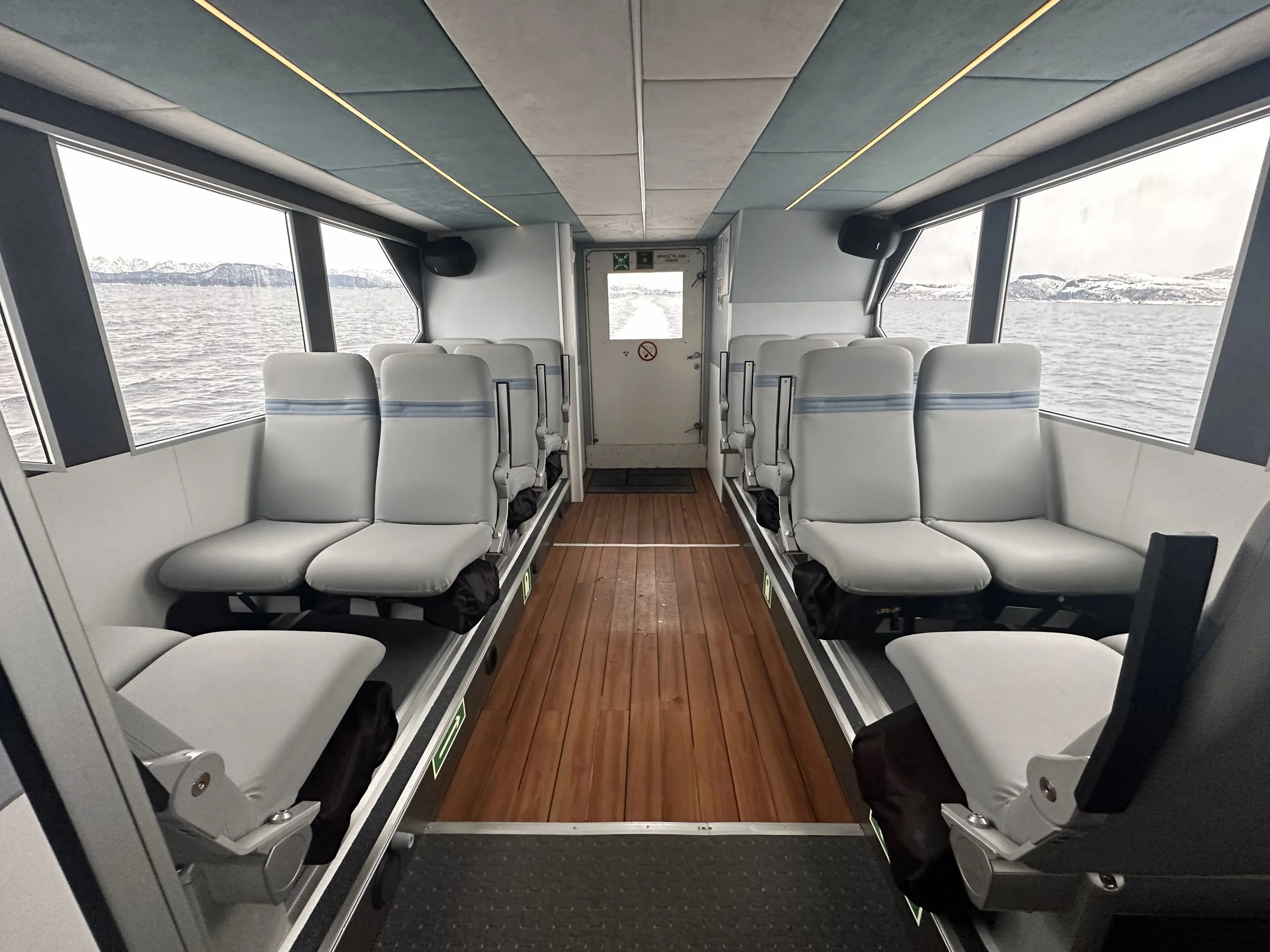 Inside of a boat with rows of white seats facing each other, large windows on both sides showing water and mountains outside, and a wooden floor in the center.